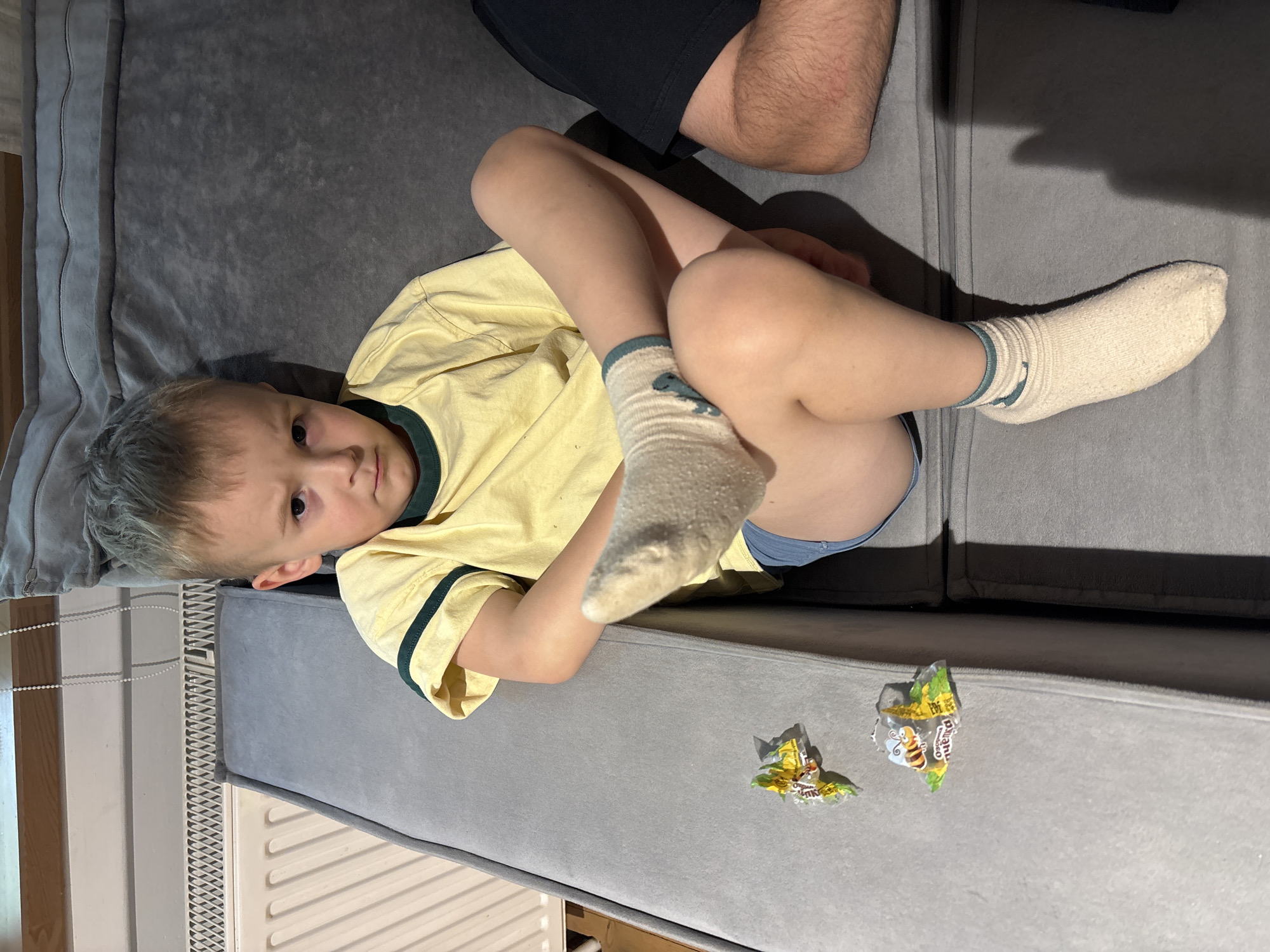 A young boy, appearing to be a child, lies on his side on a grey upholstered sofa in an indoor setting, likely a living room, in Chaikovsky, Russia. He is dressed in a yellow t-shirt with a dark green collar and sleeve trim, light blue underwear or shorts, and light-colored (possibly white or cream) socks featuring a blue-green design near the cuff, which resembles a stylized wave or abstract creature. His knees are bent, and he looks directly at the camera with a neutral, slightly wary expression.
A portion of an adult's leg or arm, covered in dark fabric, is visible in the upper right corner of the frame, suggesting another person is nearby. On the sofa, near the boy's lower body, are two crumpled candy wrappers, predominantly green and yellow. One wrapper clearly displays a cartoon bee and partial text including "BEE" and "Original," with hints of Cyrillic script that likely translate to "little bee" (пчелка) and "honey" (мед).
The lighting suggests daytime, with light coming from a source outside the frame or from a window. Behind the sofa, a window with white vertical blinds or a chain pull mechanism, and a white radiator or vent are visible, confirming the indoor environment. The image is taken from a slightly elevated, angled perspective.