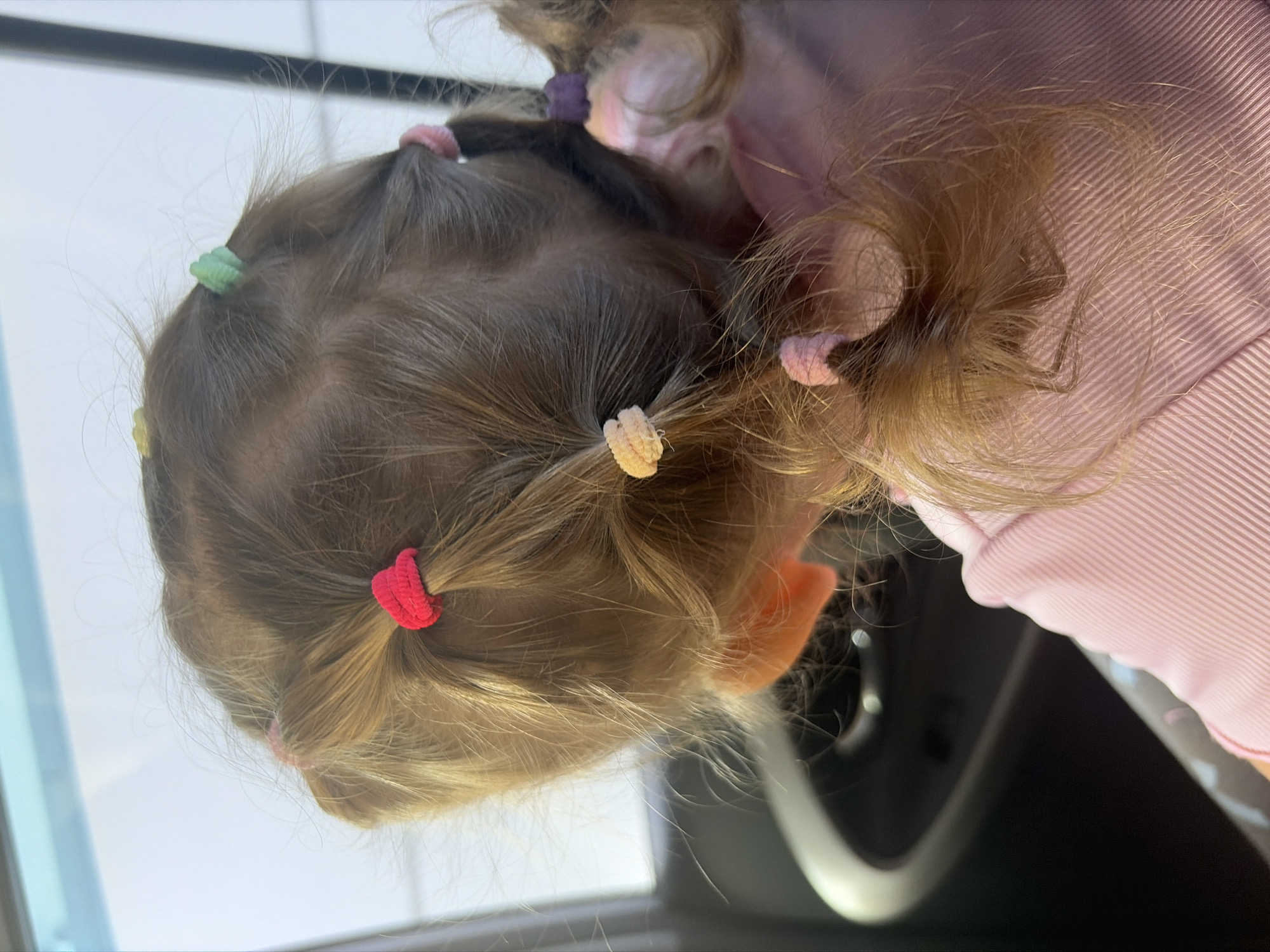 The image shows the back of a young child's head in a car. The child has light brown hair styled in multiple small ponytails secured with colorful elastic bands. The hair is a mix of straight and slightly wavy strands, with some flyaways. The child is wearing a pink ribbed shirt. The background shows a car window with a blurry view of what appears to be an outdoor environment, possibly a street or sky. The interior of the car is visible with a dark-colored door panel. There is no information in the image to suggest the location is Rostov-on-Don, Russia.