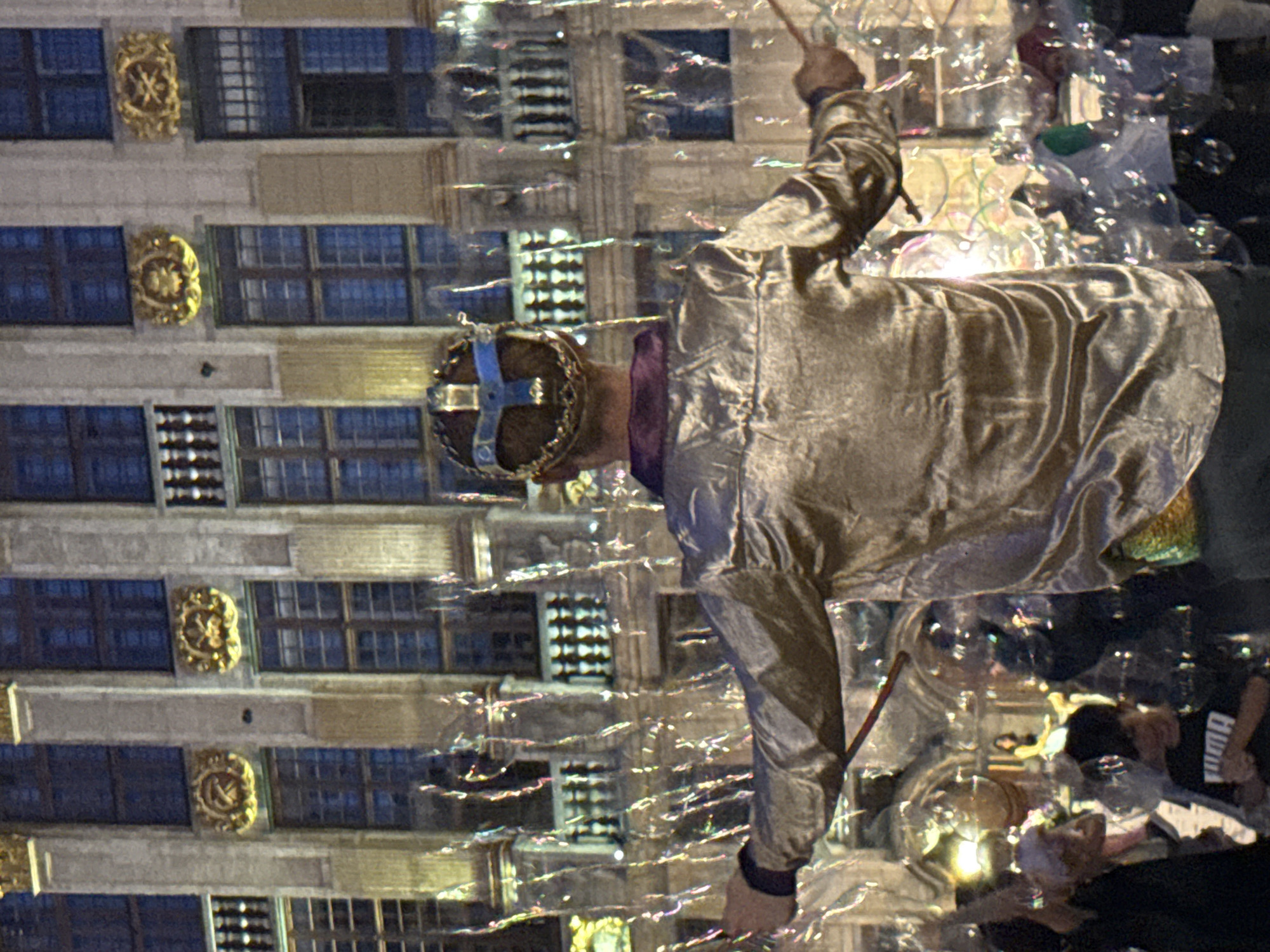 In front of the ornate facade of the Grand Place in Brussels, Belgium, a person wearing a regal costume including a crown and a shimmering gold shirt is surrounded by a profusion of iridescent soap bubbles. The person is holding a stick, suggesting an action related to bubble creation or manipulation. In the background, the intricate architecture of the historic buildings, characterized by arched windows and golden embellishments, is visible. Several other people are present in the foreground and midground, some obscured by the bubbles, contributing to a festive and public atmosphere. The lighting suggests it is evening or dusk, with artificial lights illuminating the scene and reflecting off the bubbles and the surrounding structures.