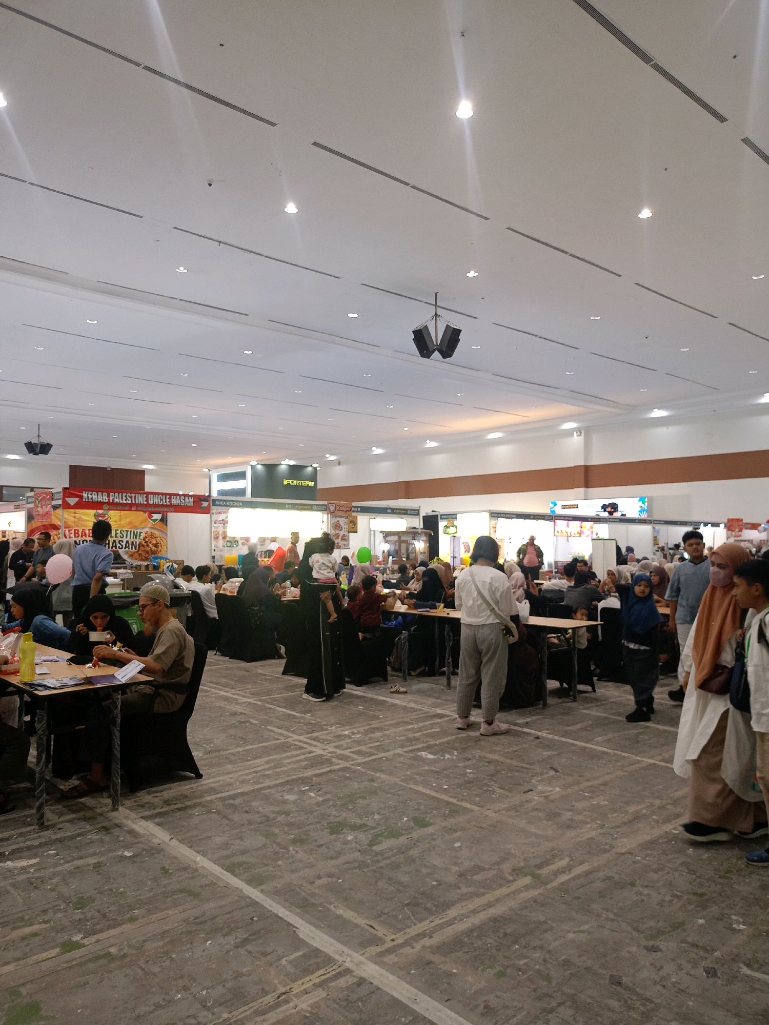The image depicts an indoor marketplace or exhibition hall, likely in Central Jakarta, Indonesia, filled with people. The scene is bustling with activity, with individuals and groups engaged in various activities.
In the foreground, several people are seated at tables, appearing to be eating or conversing. One individual in a brown shirt and a white cap is actively eating. To the left, a person in a black headscarf is also seated at a table.
Further back, a crowd of people mills about. Some are standing, while others are walking through the aisles. Several food stalls or vendor booths are visible, adorned with signage, including "KEBAB PALESTINE UNCLE HASAN" and "SHEA KITCHEN." Balloons, one pink and one green, are also present, adding a festive element to the environment.
The architecture of the space is characterized by a high, white ceiling with integrated lighting and ventilation systems. Speakers are suspended from the ceiling in some areas. The floor is a worn, concrete surface marked with white lines, suggesting it might be a repurposed venue such as a convention center or exhibition hall. The overall impression is one of a lively community event.