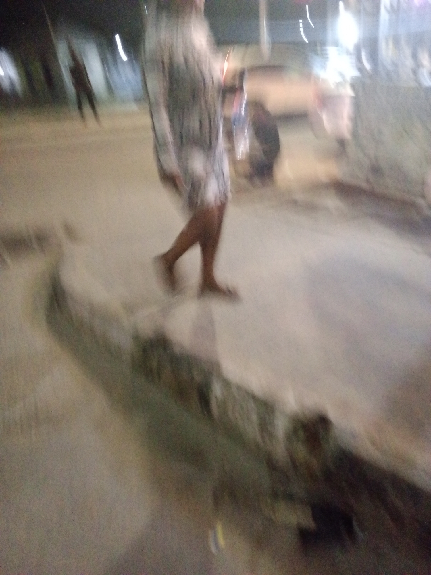 The image depicts a blurred scene at night, likely in Ado, Nigeria. A person in a patterned dress walks on a concrete surface, possibly a sidewalk or road, in the foreground. A second person is visible further away, walking in the same direction. A vehicle is visible in the background, along with some street lights. The overall scene is poorly lit and blurred, suggesting motion or a low-quality image capture. The foreground appears to show a curb or edge, indicating the boundary of the walking area. The environment seems urban, but details are indistinct due to the blur.