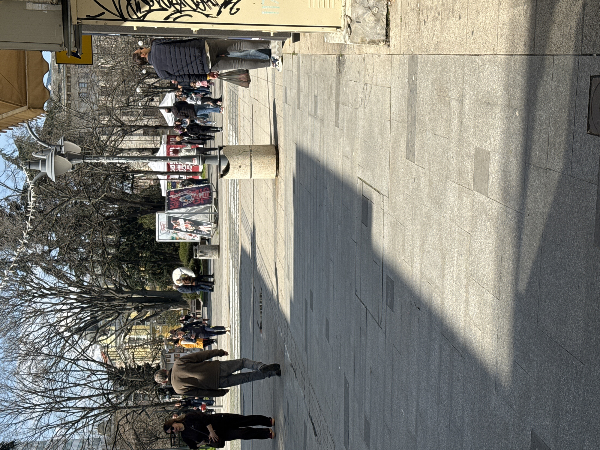 This photograph depicts a street scene in Burgas, Bulgaria, likely a pedestrian zone in the city center. The image is taken at ground level. The street is paved with grey stone and brick. The area is populated with people of varying ages wearing casual clothing, walking. Bare trees line the street along with streetlights and billboards displaying advertisements. A stone trash receptacle is visible. The buildings appear to be shops.