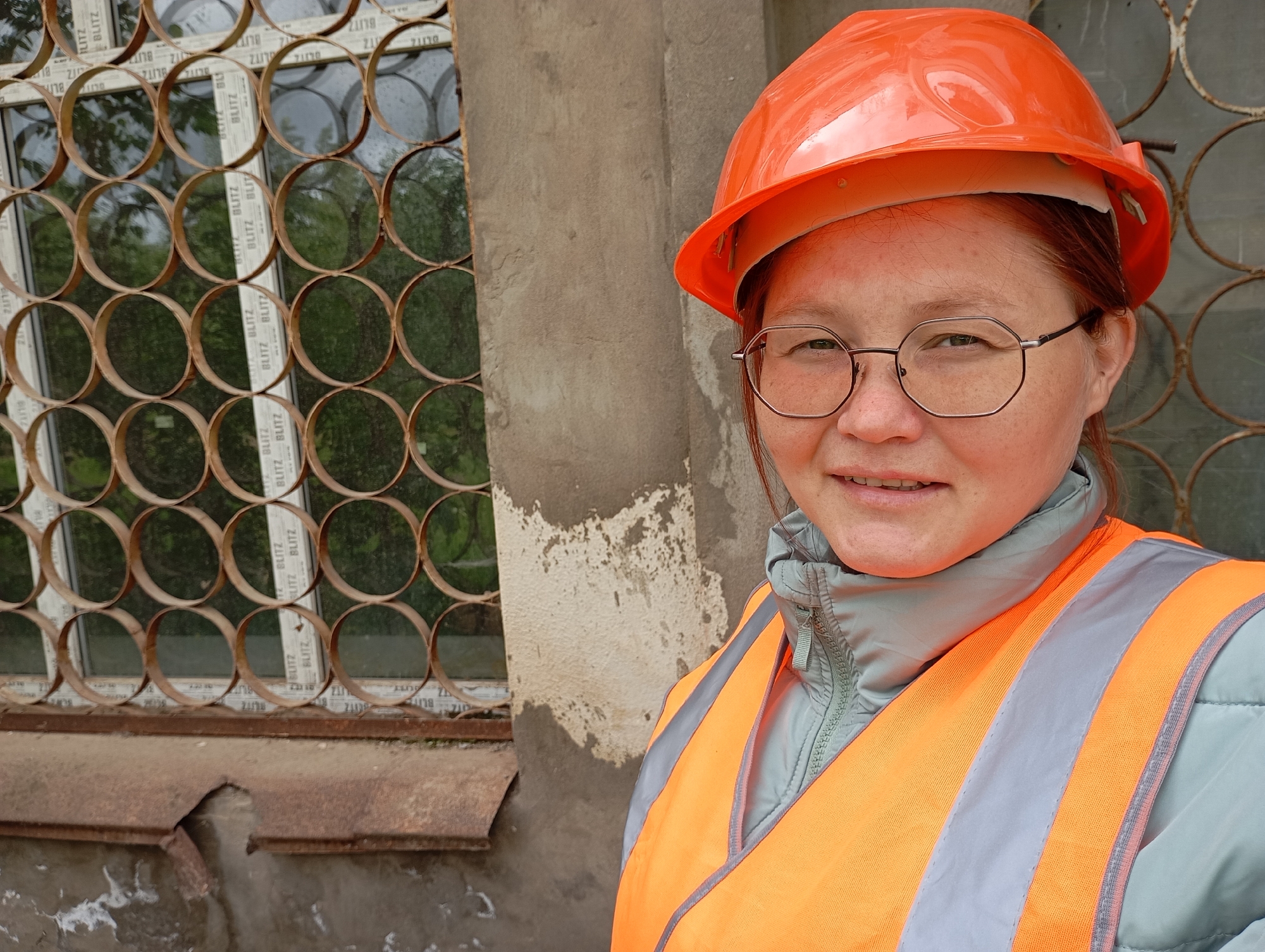 A close-up, eye-level portrait features a woman wearing an orange hard hat and an orange high-visibility safety vest over a light blue puffer jacket. She has reddish-brown hair, octagonal-framed glasses, and a slight, friendly smile, with freckles visible on her face.
The background consists of a weathered concrete or stucco wall on the right, showing signs of peeling paint or wear. To the left, there is a window covered by a decorative metal grate made of numerous interconnected circular elements, which appear somewhat rusted. Through the windowpanes, reflections of green foliage suggest an outdoor setting with trees or bushes. The white window frames clearly display the brand name "BLITZ" repeatedly, indicating the type of window profiles used. Below the window, a damaged and rusted metal sill is visible.
The woman's attire, comprising a hard hat and safety vest, indicates she is likely involved in construction, engineering, or a related profession, possibly as a worker or supervisor. The scene appears to be set outdoors during the daytime, under soft, even lighting, possibly an overcast day. The overall impression is of a practical work environment in Chaikovsky, Russia.