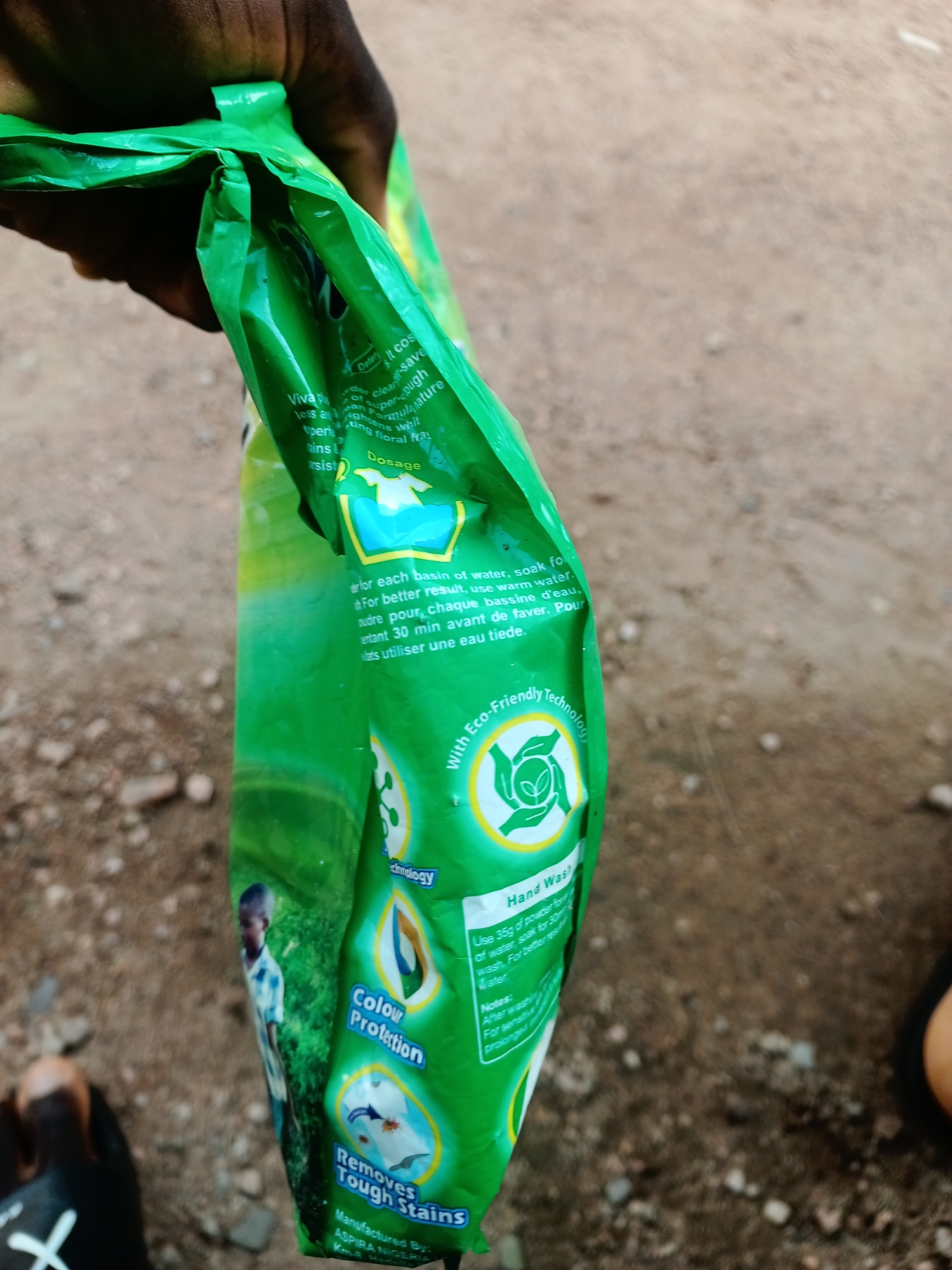 The image shows a close-up of a green plastic bag, likely containing detergent, being held by a person's hand. The bag is partially folded, revealing printed text and graphics. In the background, the ground appears to be dry and dusty, with some small pebbles visible, suggesting an outdoor setting. Part of a young boy's torso and head is visible on the left side of the bag, looking towards the left.
The text on the bag includes instructions and product claims in English and French. Notable text includes "Dosage", "Hand Wash", "Colour Protection", "Removes Tough Stains", "With Eco-Friendly Technology", and "Manufactured By ASPIRA NIGERIA". The bag also features graphics, including a stylized leaf within a circle and an image of a shirt with a stain being removed.
The lighting suggests it could be daytime. The overall scene appears to be casual and practical, possibly depicting someone examining or using the product. The presence of the boy and the outdoor setting with a dry ground points towards a location in a warm climate. The overall impression is one of everyday life, with a focus on household cleaning.