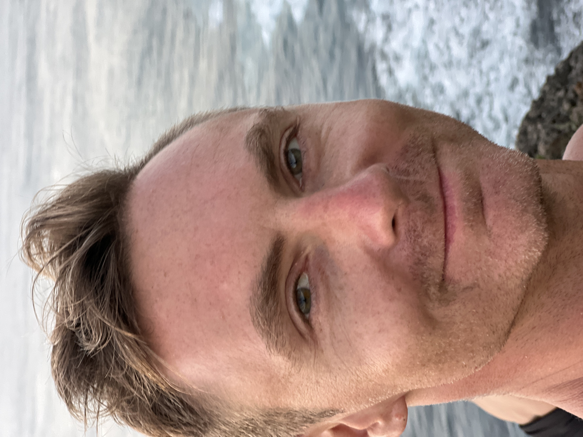 A close-up selfie shows a man's face and upper torso against a blurred background of ocean waves and sky. The man has light brown, tousled hair, fair skin with some redness on his cheeks and nose, and green eyes. He is smiling slightly, and his mouth is closed. He appears to have stubble on his chin and upper lip. The background suggests he is outdoors, possibly near a coastline, with the ocean on the right side of the frame. There is a rocky outcropping visible at the bottom right. The lighting is soft, indicating it might be daytime or dusk. The image appears to have been taken from a low angle.