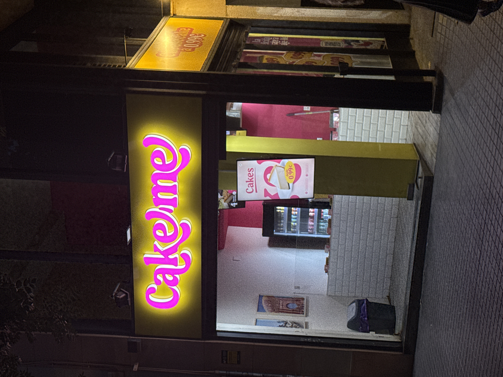The image shows the exterior of a small food establishment at night, likely a dessert shop or bakery, with a bright pink neon sign that reads "Cake me" against a yellow illuminated background. The establishment is located on a city street, with a tiled sidewalk in the foreground and a dark building facade behind it. Inside the shop, a counter is visible with white brickwork, and a small refrigerator is stocked with drinks. A promotional poster displaying a slice of cheesecake with the price "0.99€" is positioned outside the entrance. The overall impression is of a casual, modern urban business. Based on the price in euros and the style of signage, this establishment is likely in a European city, and the provided context suggests it is in Barcelona, Spain. No people are clearly visible in the image, though a portion of a person's dark clothing is visible on the far right edge, suggesting someone may be walking past.