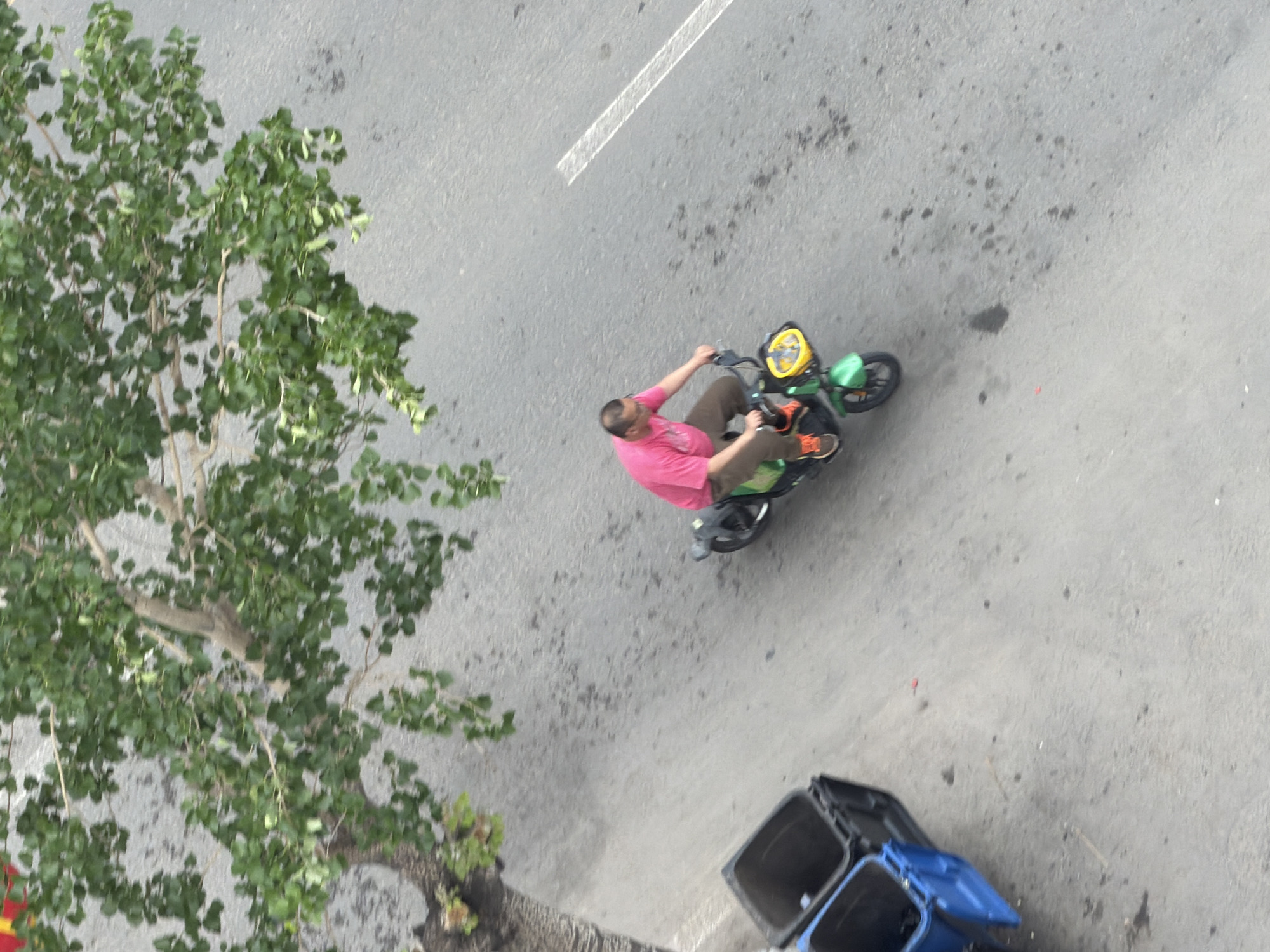 An overhead shot captures a man riding a green and yellow scooter on a paved road, likely in Tongjiang, China. The man, wearing a pink t-shirt, dark pants, and orange shoes, is positioned in the middle-right of the frame, steering the scooter. His body is angled slightly, suggesting movement.
To the left, a large tree with lush green leaves partially obscures the road. The road surface is grey asphalt, showing some wear, stains, and a clear white painted line running diagonally from the top center towards the left. In the bottom right corner, two trash cans are visible: one black and one blue. The high-angle perspective looks directly down onto the scene, offering a unique view of the street activity and surroundings. The lighting indicates it is daytime. No discernible text is visible on any object.