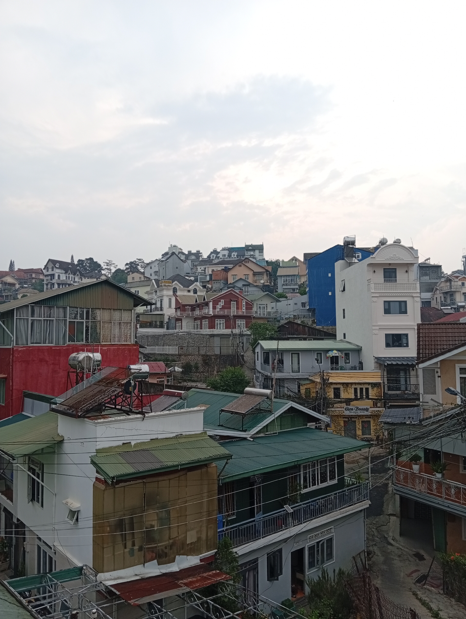 The photograph captures a dense urban scene in Da Lat, Vietnam, known for its hillside development. The view is elevated, looking across a landscape dominated by closely packed buildings of varying heights, colors, and architectural styles. The structures range from simple, utilitarian buildings to more elaborate designs.
Roofs are primarily corrugated metal, exhibiting shades of green, red, and brown. Many buildings have solar water heaters or water tanks on their roofs. The façades are painted in a variety of colors including red, white, gray, yellow, and blue.
In the background, additional buildings rise up the hillside, gradually disappearing into the cloudy sky. Power lines crisscross the scene. The perspective is from a building overlooking this cluttered urban landscape. There are no people visible in the image.