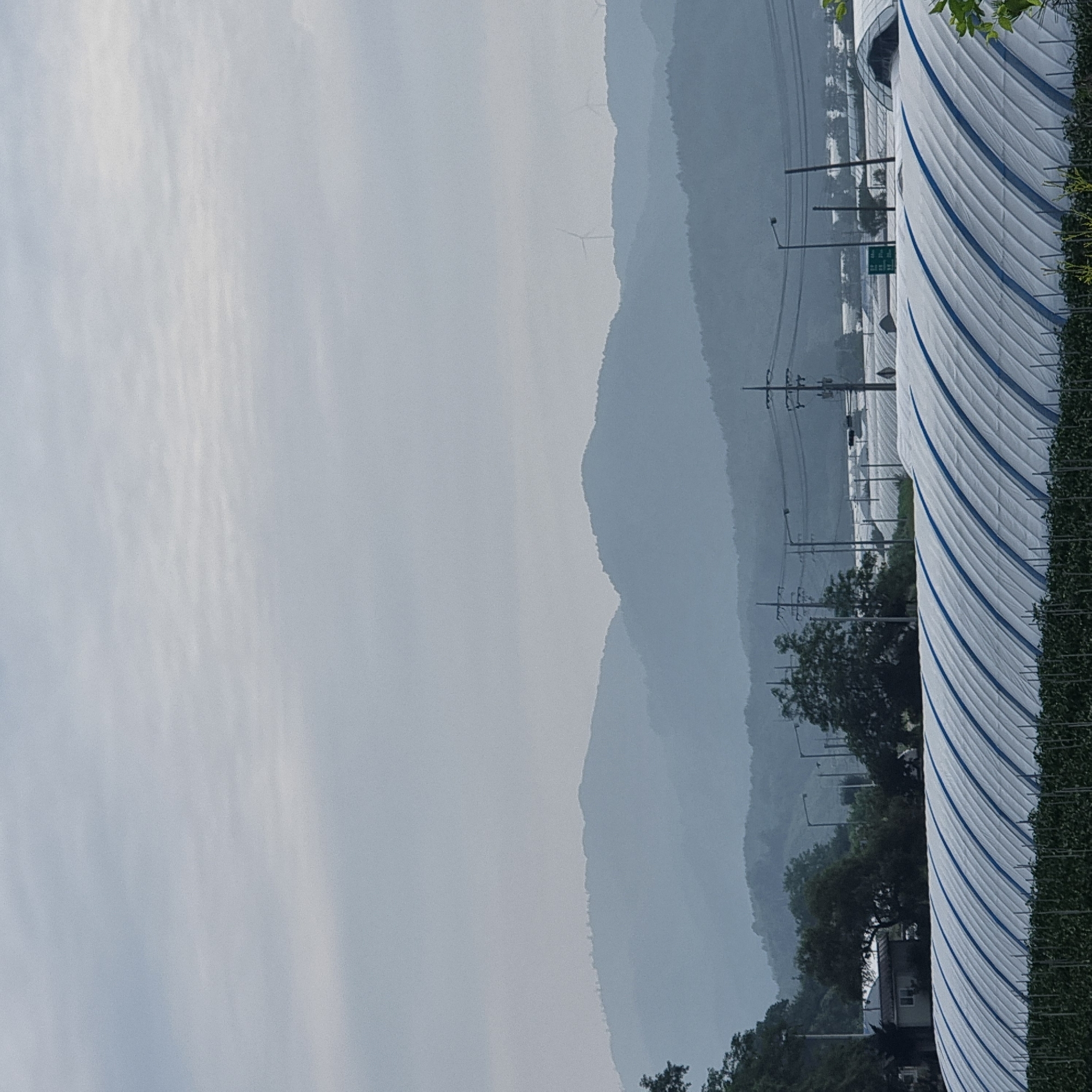 In Dunnae-myeon, South Korea, an agricultural landscape unfolds under a hazy, overcast sky. In the foreground, rows of curved greenhouses, covered in white plastic sheeting with blue trim, stretch across the frame, with the tops of plants visible below their edges. Beyond the greenhouses, utility poles and power lines punctuate the scene. In the mid-ground, a small building with a gray roof and white walls is nestled amongst trees. Further in the distance, multiple layers of mist-shrouded mountains recede towards the horizon. Wind turbines are visible atop some of the distant mountain ridges. The overall impression is one of a rural, potentially mountainous region engaged in agriculture, veiled in atmospheric haze.