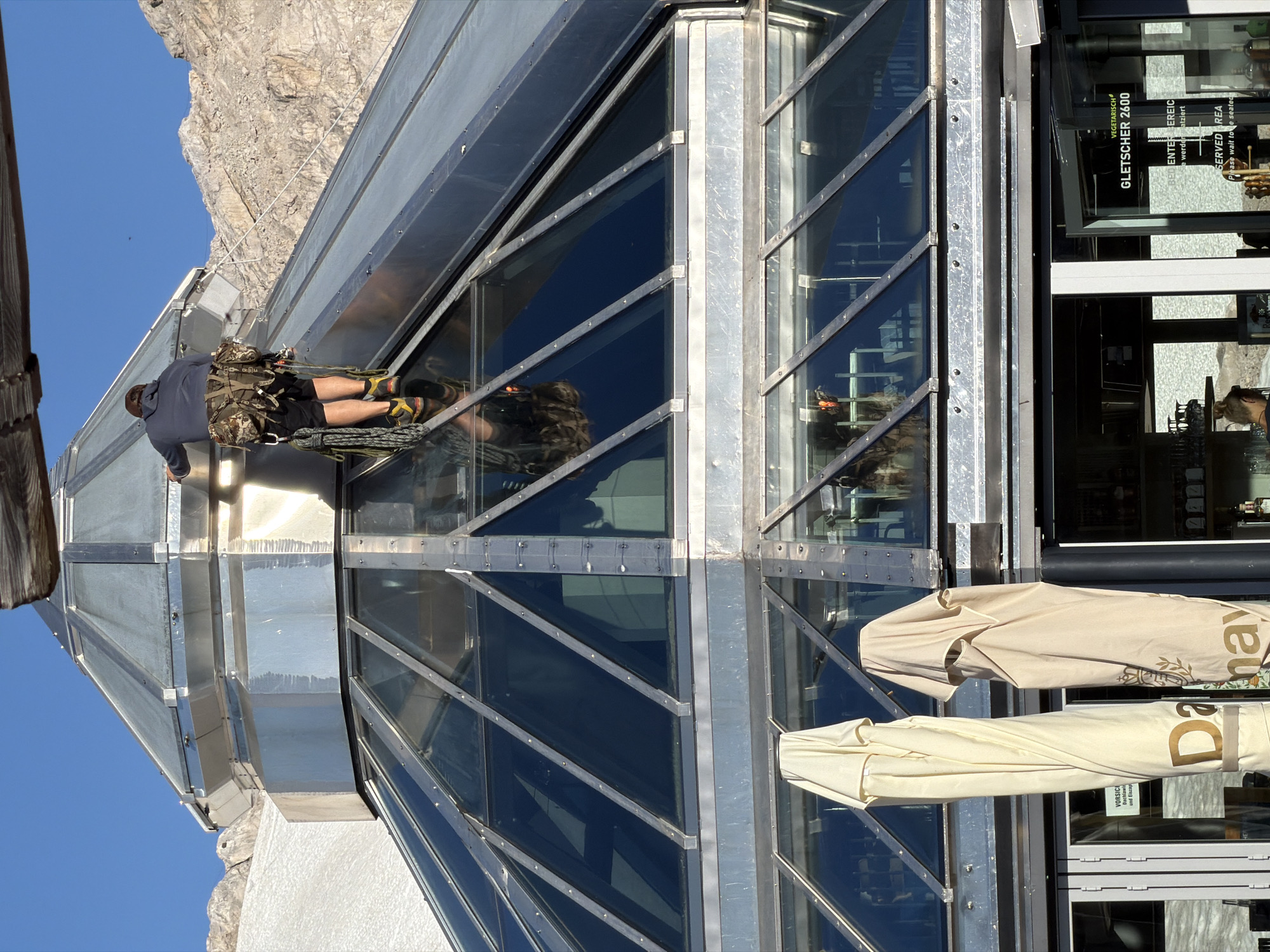 A person wearing a harness and shorts is rappelling down the side of a modern, angular building with large glass windows. The building appears to be a structure at a high altitude, indicated by the rocky mountain visible in the background and the clear blue sky. The person is secured by ropes and is positioned horizontally, facing the building. Inside the building, through the glass, shelves and other interior elements are visible. Outside the building, several large umbrellas are stacked and covered. Signs on the building include "VEGETARISCH", "GLETSCHER 2600", and "SERVED AREA", suggesting this is a restaurant or visitor center located at a glacier, likely at an elevation of 2600 meters. The scene is brightly lit, indicating daytime, and the weather appears clear and sunny. The overall impression is one of a functional building in a mountainous, possibly tourist-oriented environment, with an individual engaged in an activity that suggests maintenance or access work.