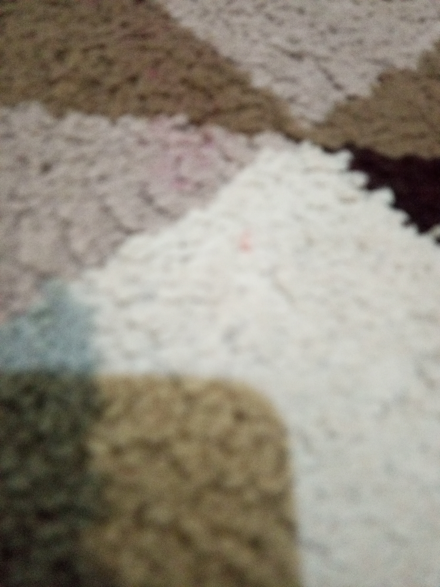 The image displays a close-up, out-of-focus view of a textured surface, likely a rug or carpet. The pattern consists of irregular shapes in muted tones of brown, beige, white, and a hint of light blue or grey. The texture appears to be fuzzy or plush. There are no discernible people, objects, actions, interactions, or environmental elements that would allow for localization within Lhokseumawe, Indonesia or any other specific location. The image is abstract due to its blurriness and lack of identifiable features.
