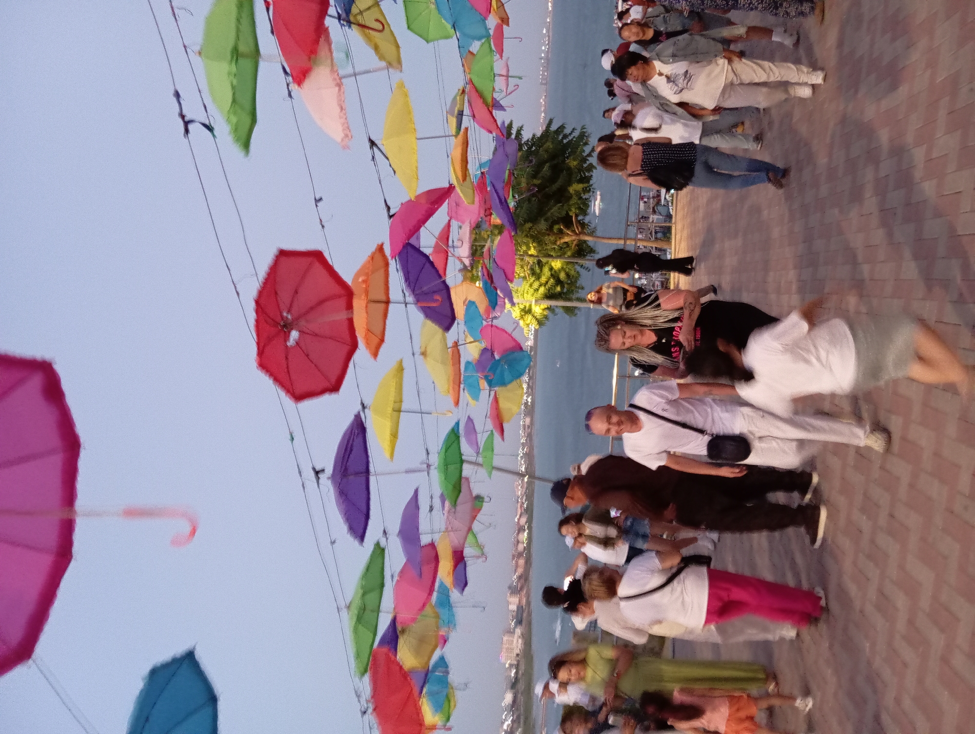 The image depicts an outdoor public space adorned with numerous colorful umbrellas suspended overhead, creating a vibrant canopy. A body of water, likely the Caspian Sea, is visible in the background, with a distant shoreline featuring buildings. Several people are walking and gathered in the foreground and midground, suggesting a recreational or event setting. The ground is paved with reddish-brown brickwork. The overall atmosphere appears to be casual and social, possibly during the evening or twilight hours given the lighting. This scene is consistent with the "Umbrella Street" installation located in Aktau, Kazakhstan, a popular attraction known for its artistic display of umbrellas above a pedestrian walkway.