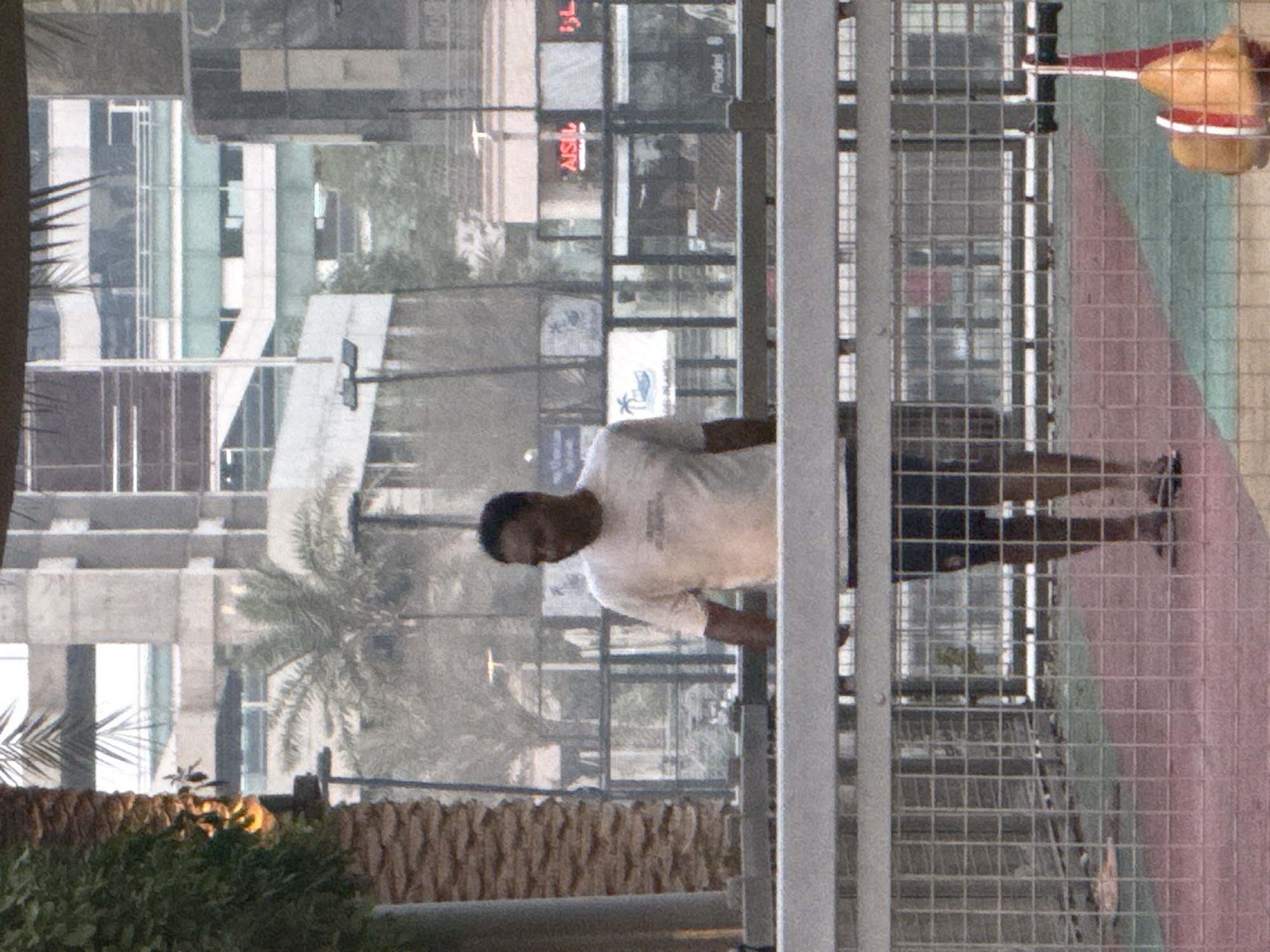 In Dubai, United Arab Emirates, a man stands in a fenced-off outdoor area, possibly a park or recreational space, with a blurry cityscape in the background. He is wearing a light-colored t-shirt and dark shorts, and his feet are bare. The area he stands in has a patterned ground with sections of red and green. Behind him, a metal fence encloses the space, and beyond that, a glass-enclosed structure or building is visible, with some signage. Palm trees and other foliage are also present in the midground. The overall atmosphere is somewhat hazy or overcast. In the foreground, a red and yellow object, possibly a piece of play equipment or signage, is partially visible.