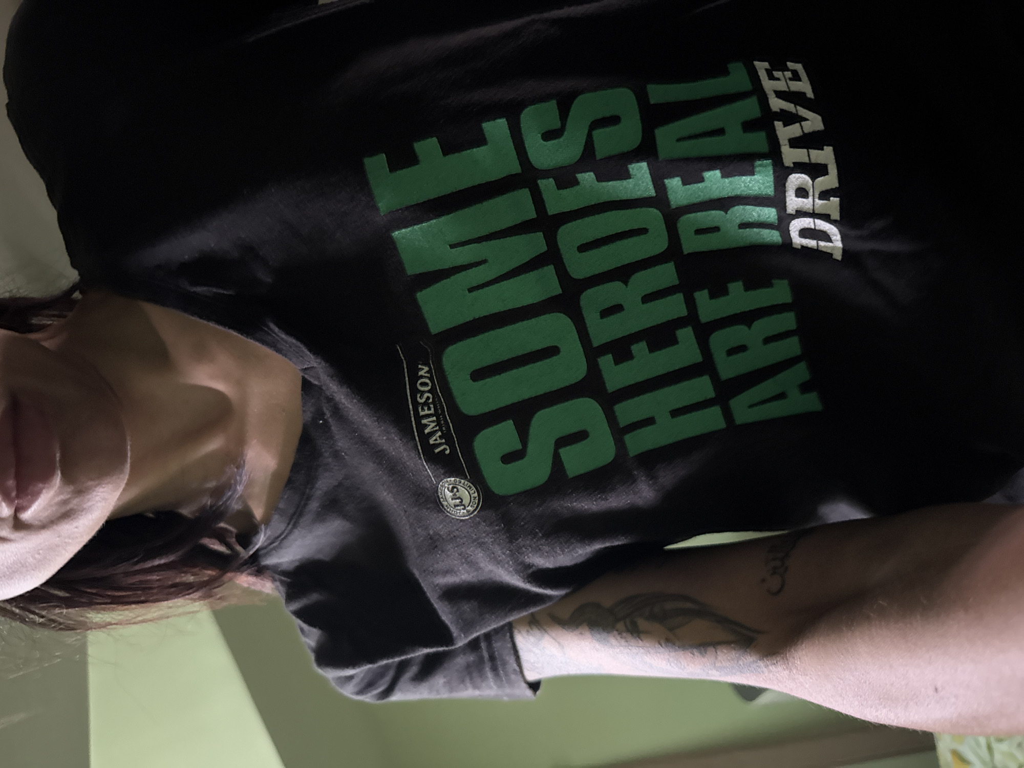 The image shows a person wearing a black t-shirt with the inscription "SOME HEROES ARE REAL DRIVE" in green and white letters, along with a "JAMESON" logo. The person has visible tattoos on their arm. The image's orientation suggests it may have been taken from a lower angle, looking up at the person. There is a light green wall or surface in the background. There is no identifiable information to place this image in the city of Pleven, Bulgaria.