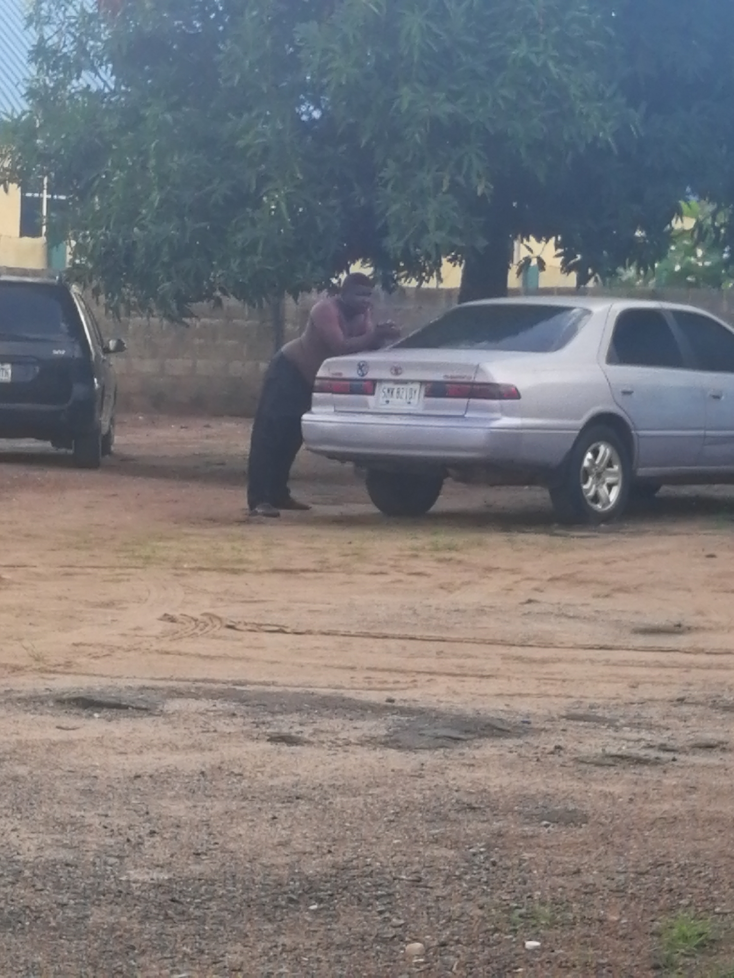 A man, shirtless and wearing dark pants, is leaning against the rear of a silver Toyota Camry parked on a dirt lot. The man's right arm is extended, and he appears to be resting his head on his arm. He is positioned to the right of the car's bumper. To the left of the Camry, a dark SUV is partially visible. Behind the vehicles, a building with yellow walls and a white-framed window can be seen, along with a large tree with green foliage. The ground is a mix of dirt and gravel, with tire tracks visible in the dirt. The scene appears to be in Ajasse Ipo, Nigeria.