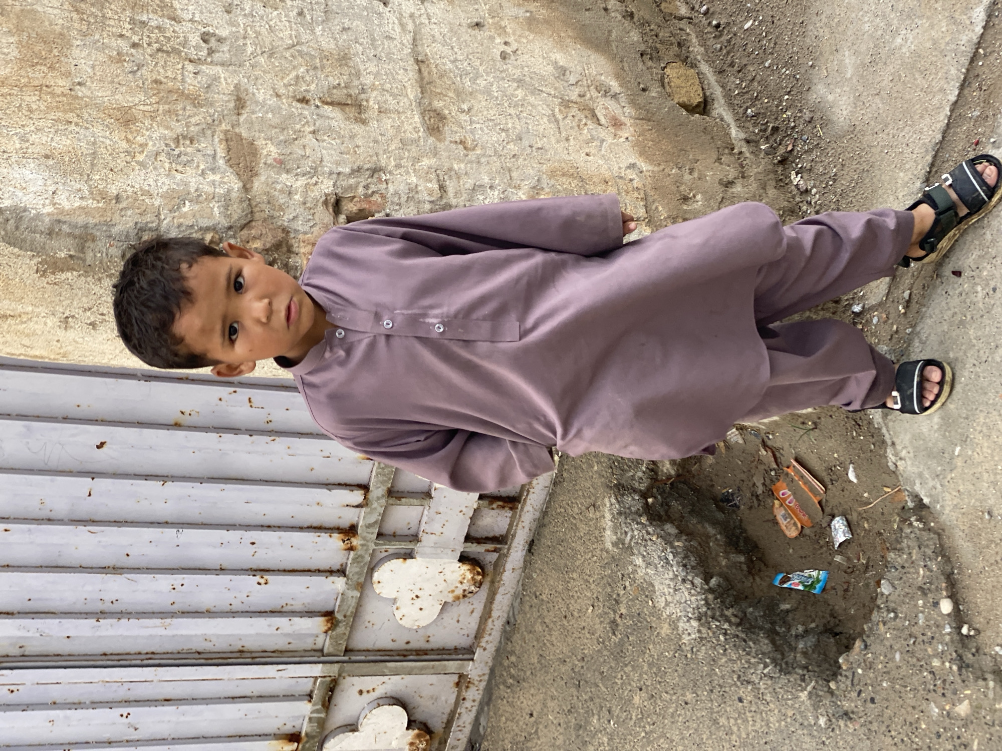 An image taken in Kandahar, Afghanistan, depicts a young male child standing outdoors, centered in the frame. The child, appearing to be between 4-6 years old, has dark hair and wears a long-sleeved, collared purple shalwar kameez with three visible white buttons on the placket and black sandals with white accents and Velcro straps. His left hand is in his pocket, and there is visible dirt on his face below both eyes. He gazes directly at the viewer with a neutral expression.
To the child's left is a light grey corrugated metal gate, heavily rusted with numerous brown spots, featuring two white clover-shaped decorative elements along its lower horizontal bar. To his right is a weathered, light brown textured wall, likely constructed from bricks or stones, exhibiting crumbling plaster and exposed material. The ground beneath the child is uneven concrete or asphalt. A noticeable depression in the ground to the child's left (viewer's right) contains several pieces of debris, including a blue and white wrapper and two orange-colored wrappers. The overall environment appears worn and urban.