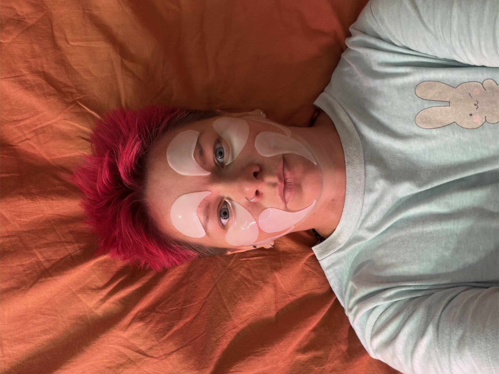 A person with bright pink, spiky hair lies on a rumpled orange surface. They are wearing a light blue t-shirt with a cartoon bunny printed on the left side of the chest. The person's face is partially covered with white under-eye patches, arranged in a symmetrical pattern around their eyes and cheeks. Their eyes are open and looking directly at the camera. The person has a nose ring and appears to be a woman. The orange surface appears to be fabric, possibly a blanket or bedsheet. The setting is indoors, and there are no discernible landmarks or environmental features that would indicate a specific location in Chaikovsky, Russia.