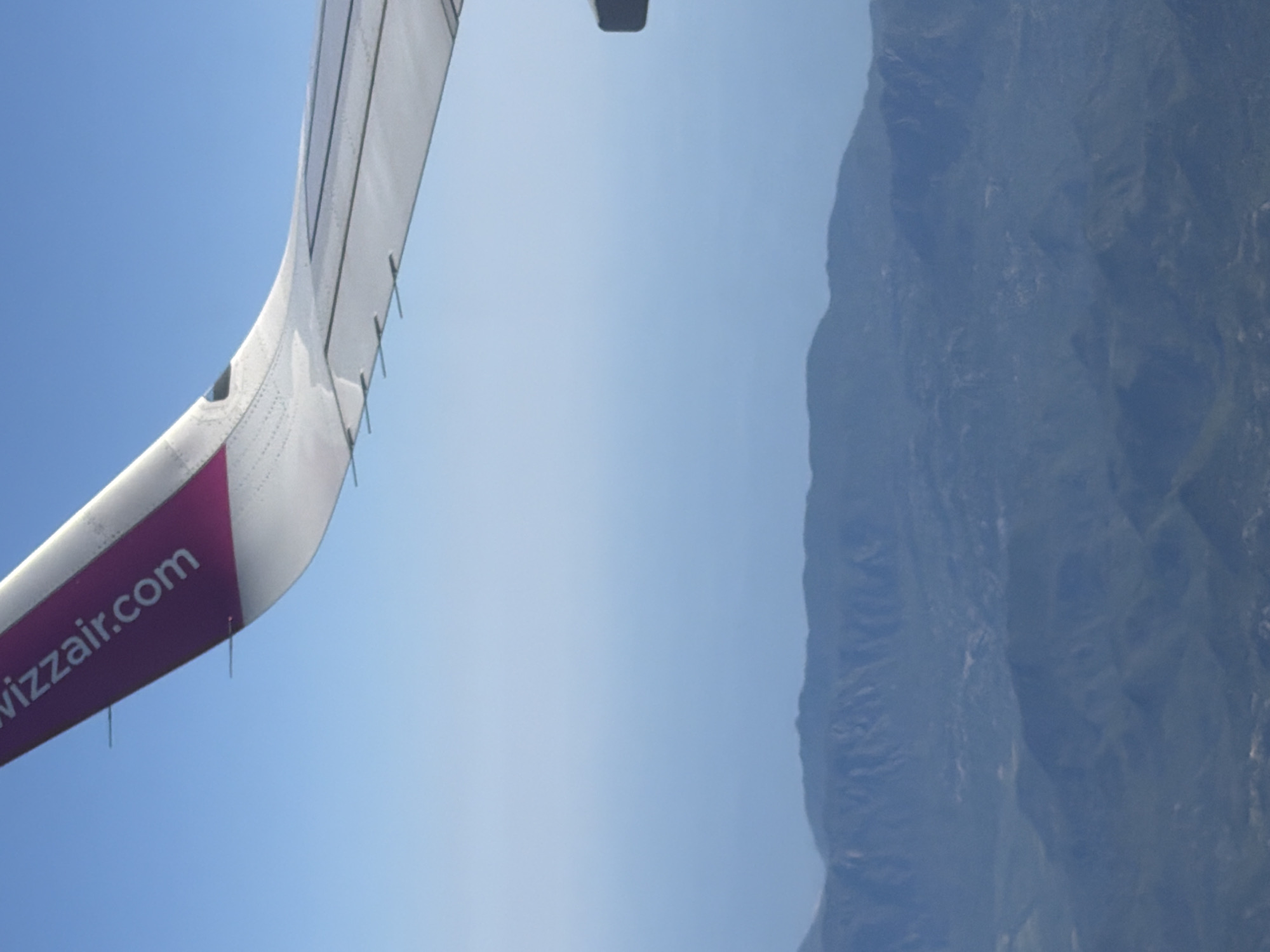An aerial view shows a portion of an airplane wing with the text "wizzair.com" printed on a purple section. The wing is angled upwards and to the right, with the clear blue sky filling the background. Below the wing, a hazy, mountainous landscape is visible. The mountains appear layered and are covered in dark green vegetation, suggesting a natural, undeveloped area. The image's perspective indicates the photograph was taken from inside the airplane. The location of Donja Cijevna, Montenegro, is not visually identifiable from the provided image.