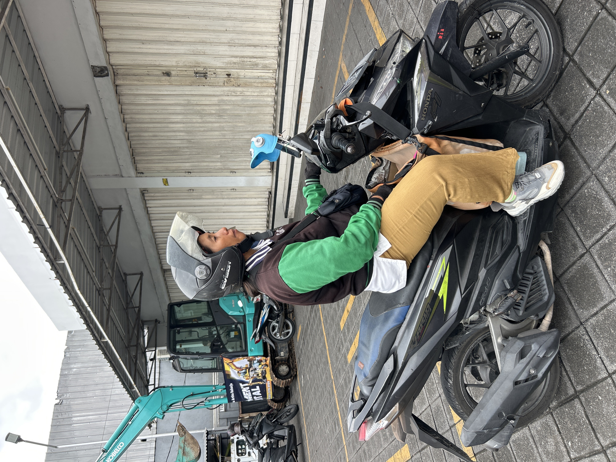 A woman wearing a helmet, a brown and green jacket over a white shirt, and tan pants, sits on a black Honda scooter. She is holding the handlebars, looking to her right. The scooter is parked on a paved area marked with yellow lines. Behind the scooter, a teal excavator is visible, along with a white building with corrugated metal siding and large roll-up doors. The scene appears to be an outdoor area, possibly a construction site or commercial space, in North Kuta, Indonesia, given the presence of construction equipment and a typical Indonesian building style.