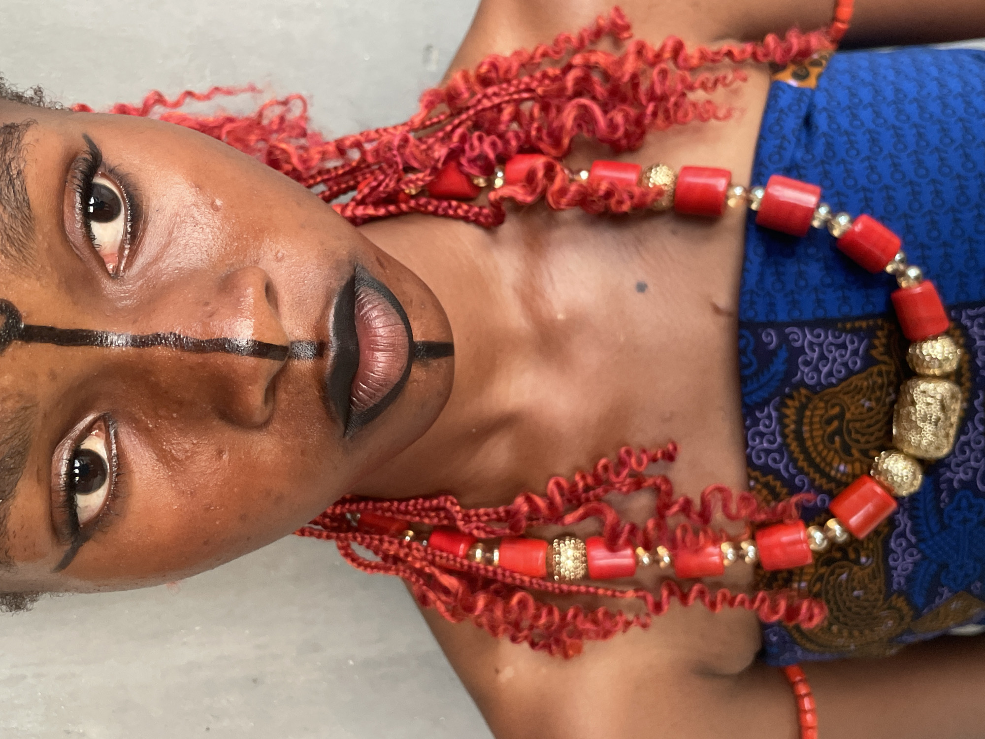 A close-up portrait captures an individual with dark skin, turned sideways, looking directly at the camera. Their face is strikingly adorned with black artistic or ceremonial paint: a prominent horizontal line crosses the bridge of the nose and extends onto the cheeks, intersected by a vertical line that runs from the forehead, down the nose, through the lips, and onto the chin.
The person's eyes feature dark winged eyeliner and mascara, while their eyebrows are neatly defined. Their lips are painted with a dark lipstick, possibly with a subtle lighter tone in the center. The skin appears luminous, reflecting light.
Around the neck and shoulders, the individual wears elaborate red beaded necklaces, accented with textured gold spherical beads and smaller gold spacers. These vibrant red beads are complemented by matching red, curly hair or intricately styled extensions that frame the face and fall across the shoulders.
On the upper right side of the frame, a blue fabric with a detailed, patterned design, likely a traditional African print, is visible, covering part of the person's torso. The background is a plain, light grey surface, emphasizing the subject.
The image suggests a deliberate pose, possibly for a cultural display, artistic project, or fashion statement, highlighting rich adornments and ceremonial face paint. The direct gaze creates an engaging connection with the viewer. There is no visible text in the image.