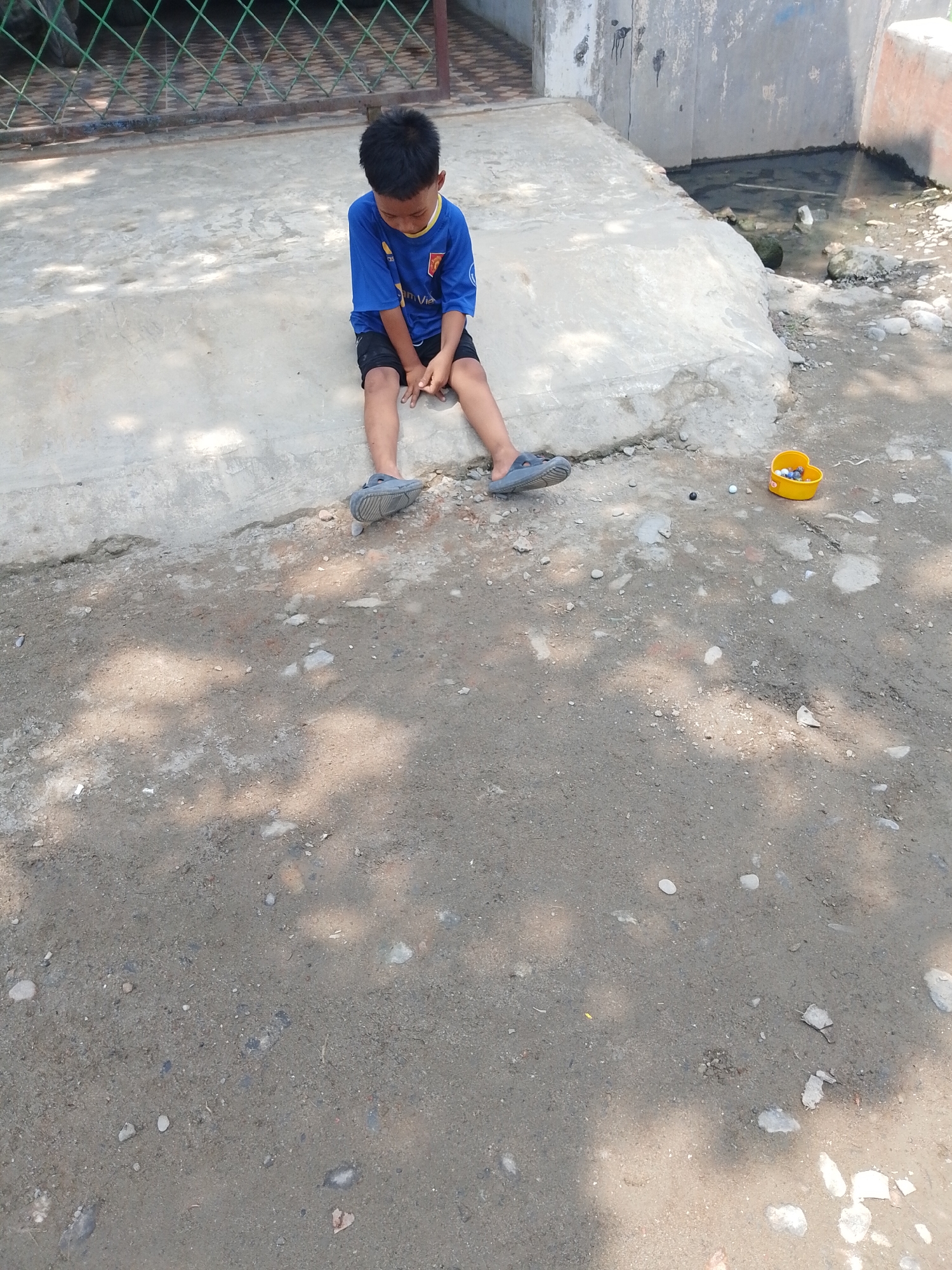 A young boy, appearing to be of Southeast Asian descent, sits on a concrete ledge outdoors. He is wearing a blue t-shirt with yellow trim and black shorts, and grey sandals. The boy is looking down with his hands clasped in his lap. The environment appears to be a somewhat unkempt urban area, possibly in the city of Hamparan Perak, Indonesia, judging by the style of the building and the presence of a drainage ditch with dark water to the right of the boy. The ground is a mixture of dirt and small stones, with shadows cast by trees or other overhead objects. A small yellow plastic container filled with marbles is visible on the ground near the boy's feet. A metal fence is in the background.
