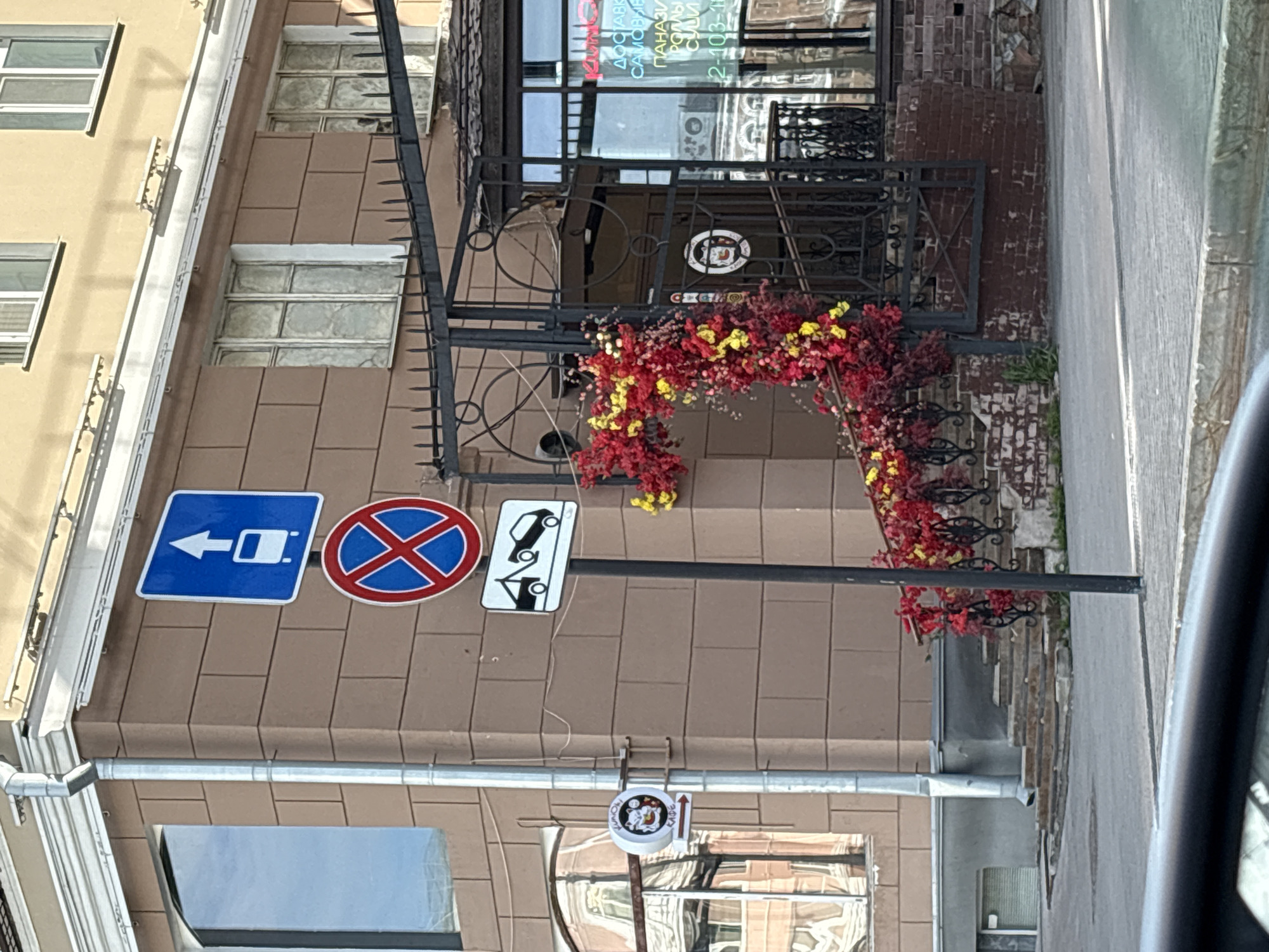 The image depicts a building facade in Perm, Russia. Three road signs are visible: a blue sign with a white car pointing left, a red circle with a blue cross indicating no stopping, and a white sign with two cars indicating a specific rule. An ornate, dark metal entrance with decorative floral arrangements leads into a business. A lower sign with a logo is present, indicating a local establishment. Reflections in the lower windows suggest a street scene. A portion of a vehicle is visible in the right side.