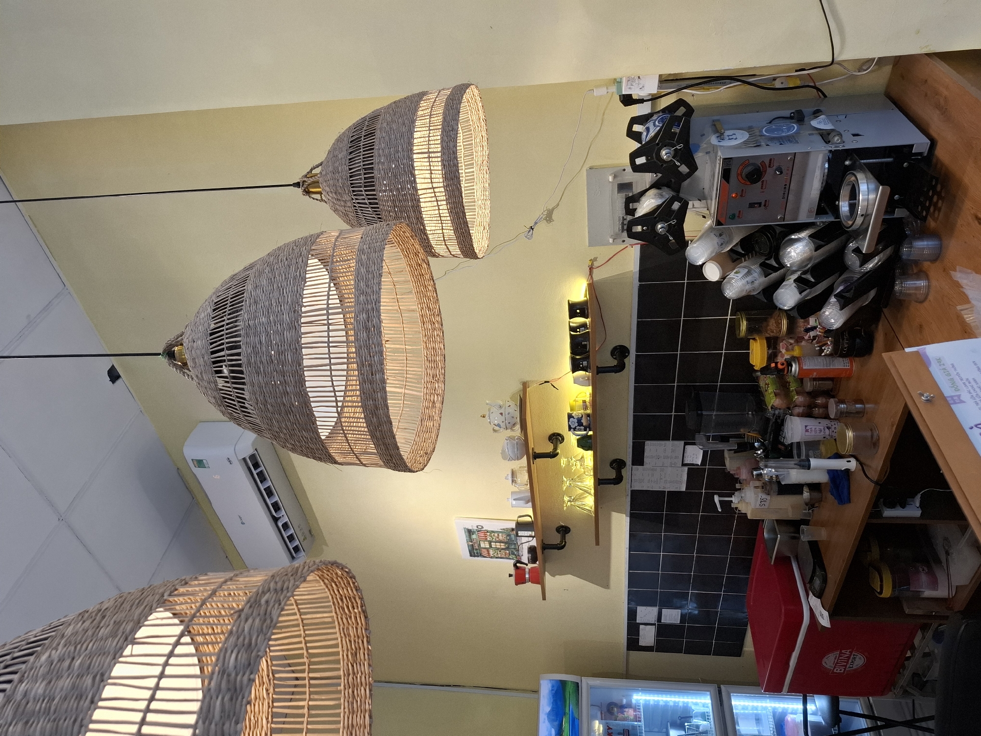 This is an interior scene of a cafe in Nha Trang, Vietnam. The image features a light yellow wall and a white ceiling. Suspended from the ceiling are three large, woven pendant lights with a natural wicker texture, casting a warm glow. On the wall, an air conditioning unit is mounted high up. Below this, two wooden shelves are installed, decorated with various items. The top shelf holds a red moka pot, a black mug, and several ceramic teacups and a teapot with a floral design. The bottom shelf displays a set of clear drinking glasses, a mug with a blue pattern, and a yellow mug.
The lower part of the image shows a wooden counter. To the left, a red cooler box with "BIVINA EXPORT" branding is visible, with a black folding chair behind it. A refrigerator with illuminated shelves is also partially visible on the far left. The counter area is equipped with a sealing machine for cups, a blender, and various containers, bottles of sauces, and other items commonly found in a cafe. The wall behind the counter is tiled with black squares. A menu or price list is displayed on a white piece of paper on the wall above the counter. The overall ambiance suggests a functional and somewhat rustic cafe setting.