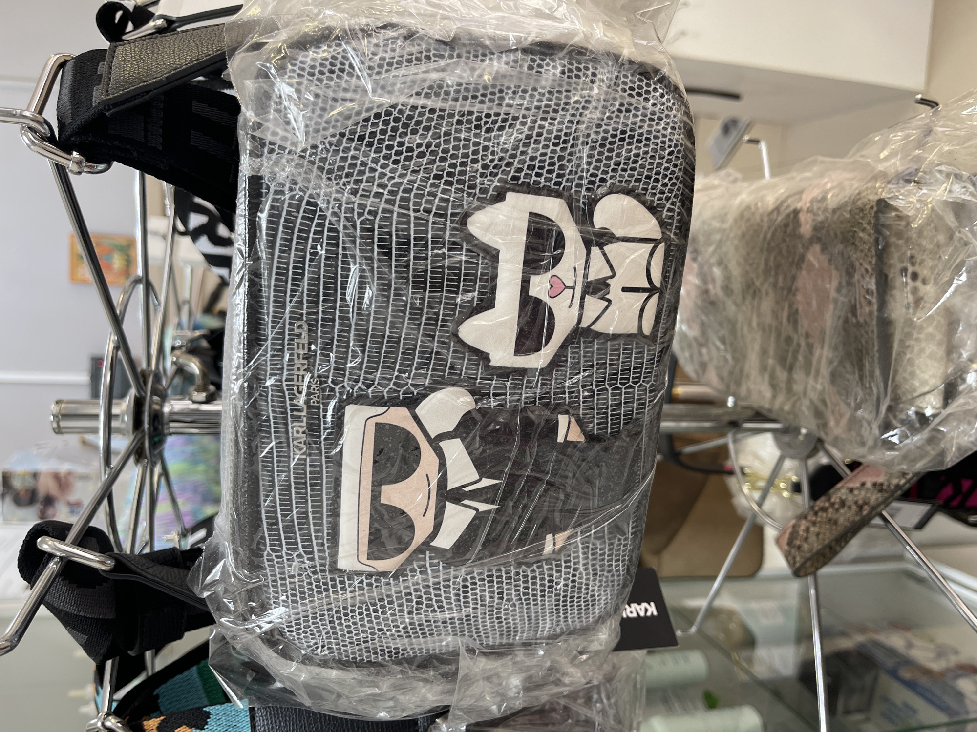 The image shows a Karl Lagerfeld bag hanging on a metal display rack. The bag is inside a clear plastic bag. The bag has a gray netting pattern and features two stylized cartoon cat logos, which appear to be versions of Lagerfeld's cat, Choupette. The rack also has other items displayed. The setting appears to be a retail environment, possibly a boutique or department store. There is no verifiable data to determine the city of Podolsk, Russia in this image.