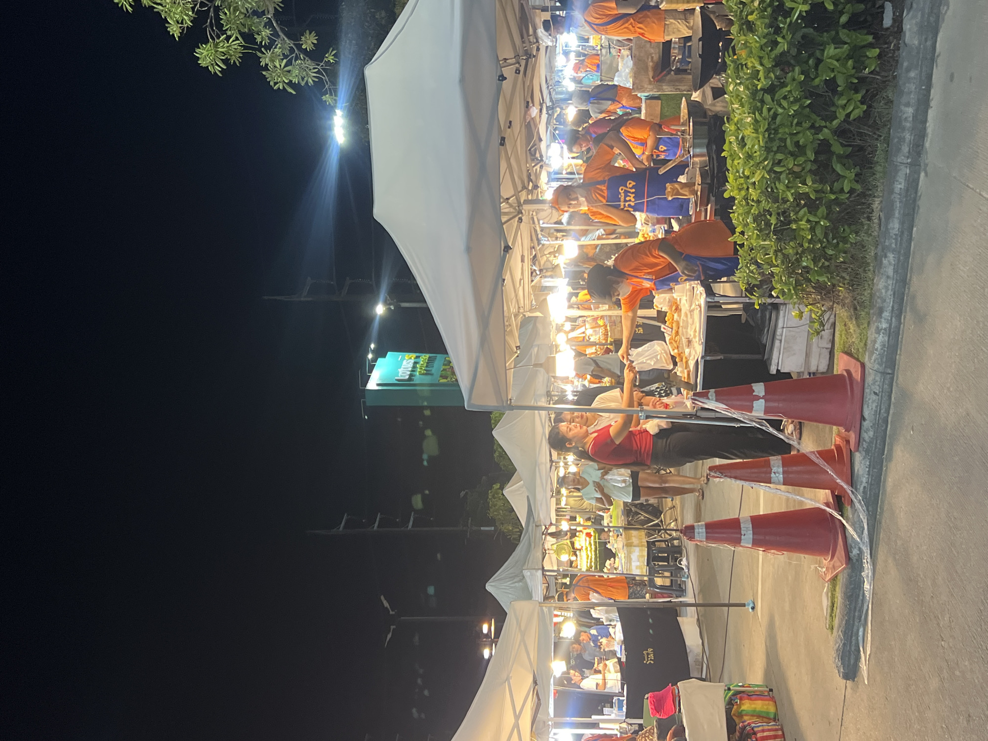 This image depicts a nighttime scene at an outdoor market in Choeng Thale, Thailand. Several stalls with white tents are set up, illuminated by artificial lights. People are actively engaged at the stalls, with some appearing to be vendors and others customers. In the foreground, three orange traffic cones are placed along a curb. The market appears to be a food market, with some stalls displaying food items and cooking equipment. The overall atmosphere suggests a lively evening market environment.