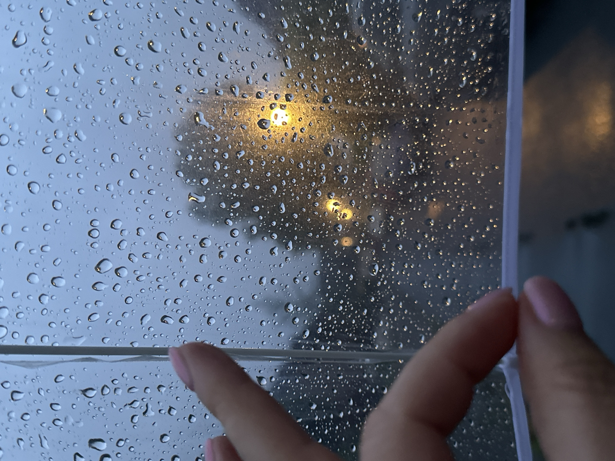 A close-up shot depicts raindrops on a transparent surface, likely an umbrella or window, obscuring a dimly lit outdoor scene. A person's hand with pink nail polish is visible in the foreground, touching the surface. In the background, out-of-focus streetlights cast a warm glow through the rain, illuminating trees and a wet road. The overall mood is somber and reflective, suggesting a rainy evening in an urban environment. The location is Krefeld, Germany.