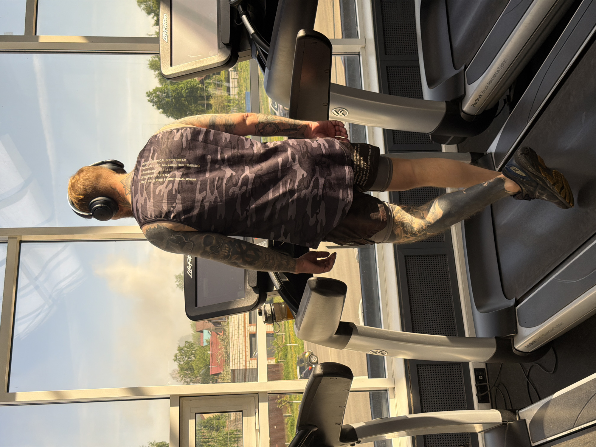 A man is exercising on a treadmill in what appears to be a gym or fitness center. The man is wearing a camouflage tank top, shorts, and sneakers, and he has headphones on. He has tattoos on his arms. The treadmill is a Life Fitness model. Large windows are behind the treadmills, offering a view of Izhevsk, Russia. Visible through the windows are buildings, trees, and possibly a car. The scene is well-lit, possibly due to the natural light streaming in from the windows.