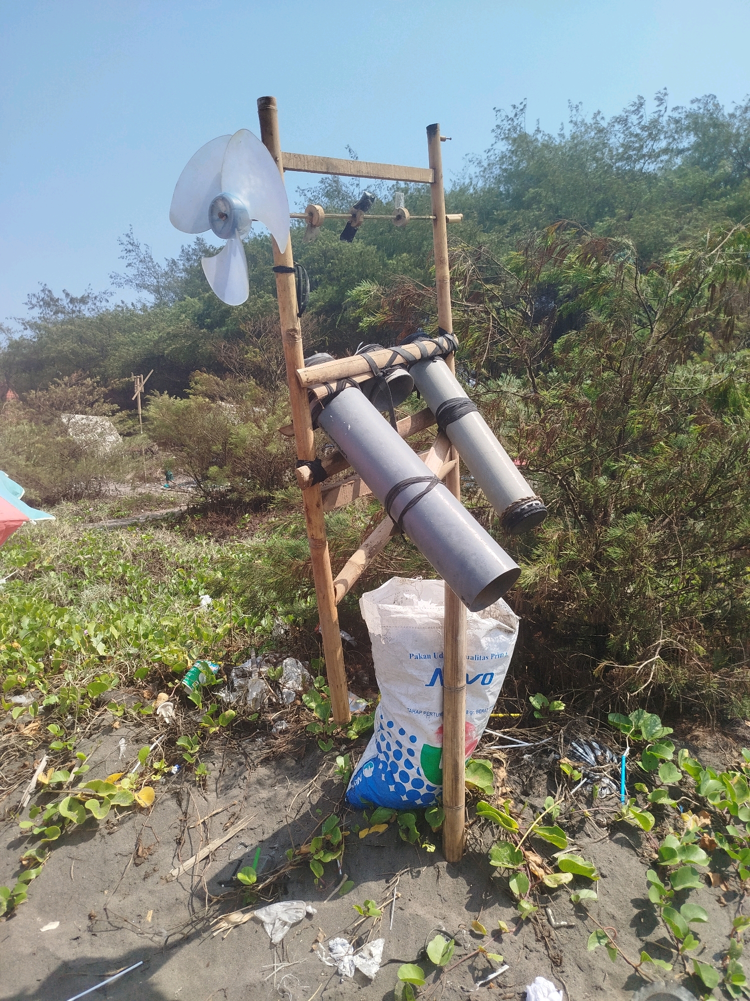 The image depicts a beach scene in Kenoyajaya, Indonesia, featuring a makeshift structure constructed from bamboo poles. This structure supports a clear plastic fan with three blades mounted on a horizontal beam, and two gray PVC pipes of unequal length are also attached to the bamboo frame, angled downwards. A white woven bag with blue lettering is placed at the base of the structure. The ground is covered in dark sand, strewn with various pieces of debris including plastic bottles, wrappers, and dried vegetation. Green ground cover plants are growing amongst the sand and debris, and in the background, a dense line of trees forms a backdrop against a clear blue sky. No people are visible in the image.