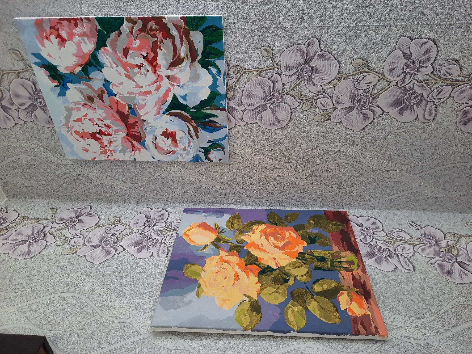 The scene depicts two paintings placed against a wall and a surface, both adorned with a floral pattern wallpaper. The upper painting showcases a collection of flowers in shades of pink, red, and white, set against a light blue background. The lower painting displays yellow and orange roses with green leaves against a blue and purple backdrop. There is a dark object partially visible in the lower left corner. The wall is decorated with embossed orchid wallpaper. The location is likely Krasnogorskoye, Russia.