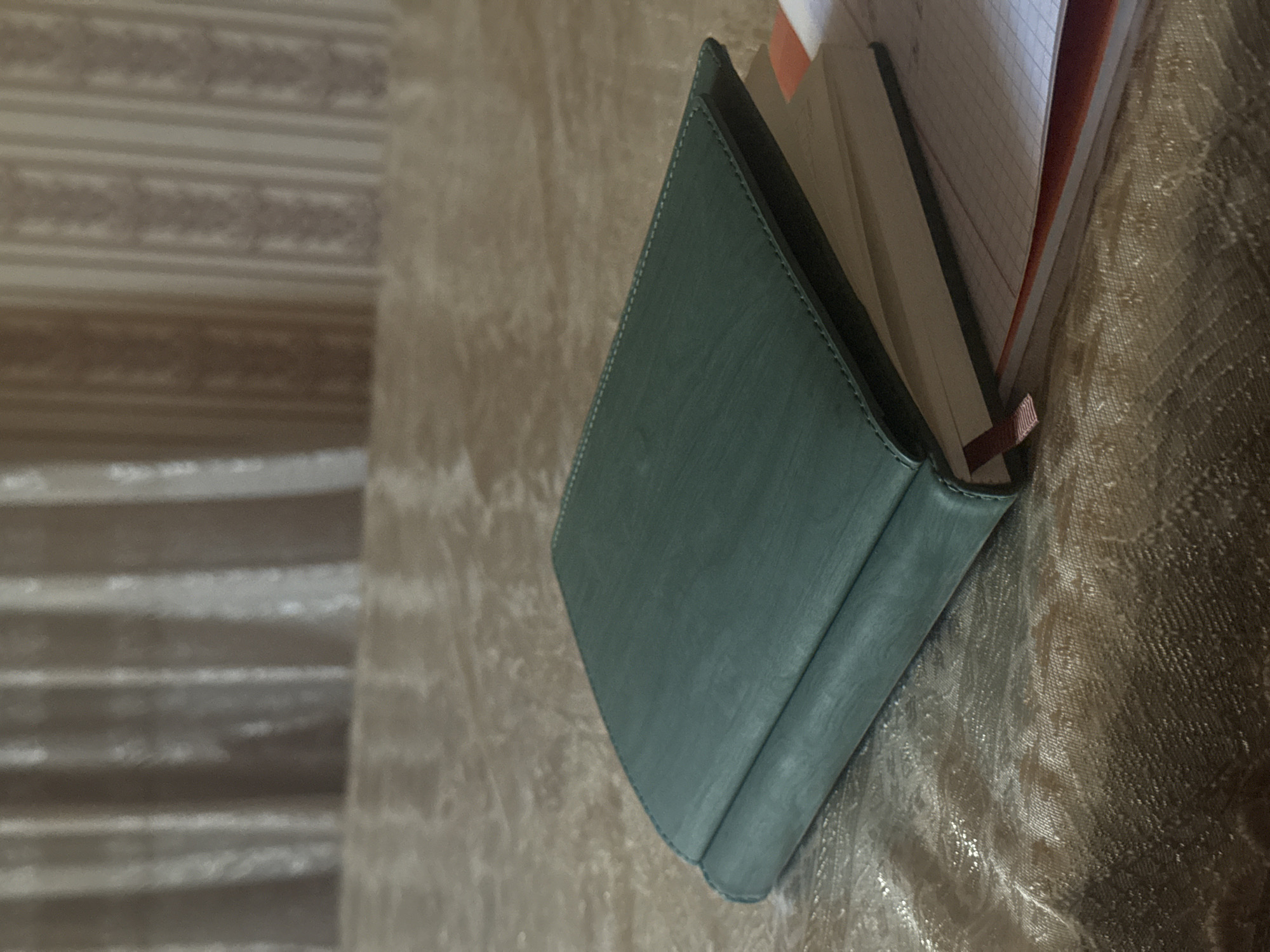 A dark green, leather-bound journal lies open on a textured, light-colored tablecloth. The journal is positioned diagonally, with its spine facing the viewer's left. The pages of the journal are visible, some appearing blank and others filled with faint grid lines. A thin, pink ribbon bookmark peeks out from between the pages. The background consists of vertically striped curtains, with the lower portion of the image focused on the tablecloth. There are no people or animals present. The lighting suggests an indoor setting, likely in Navbahor, Uzbekistan, given the contextual information.