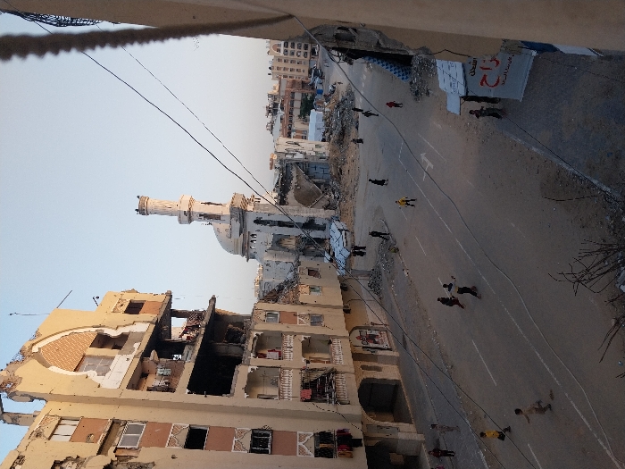 A street with destroyed buildings, including a mosque with a minaret and dome, is depicted. The buildings are heavily damaged, with exposed interiors and missing walls. Several people are walking along the street, and utility lines stretch across the scene. A sign is visible on a building in the background. The overall impression is one of destruction and aftermath.