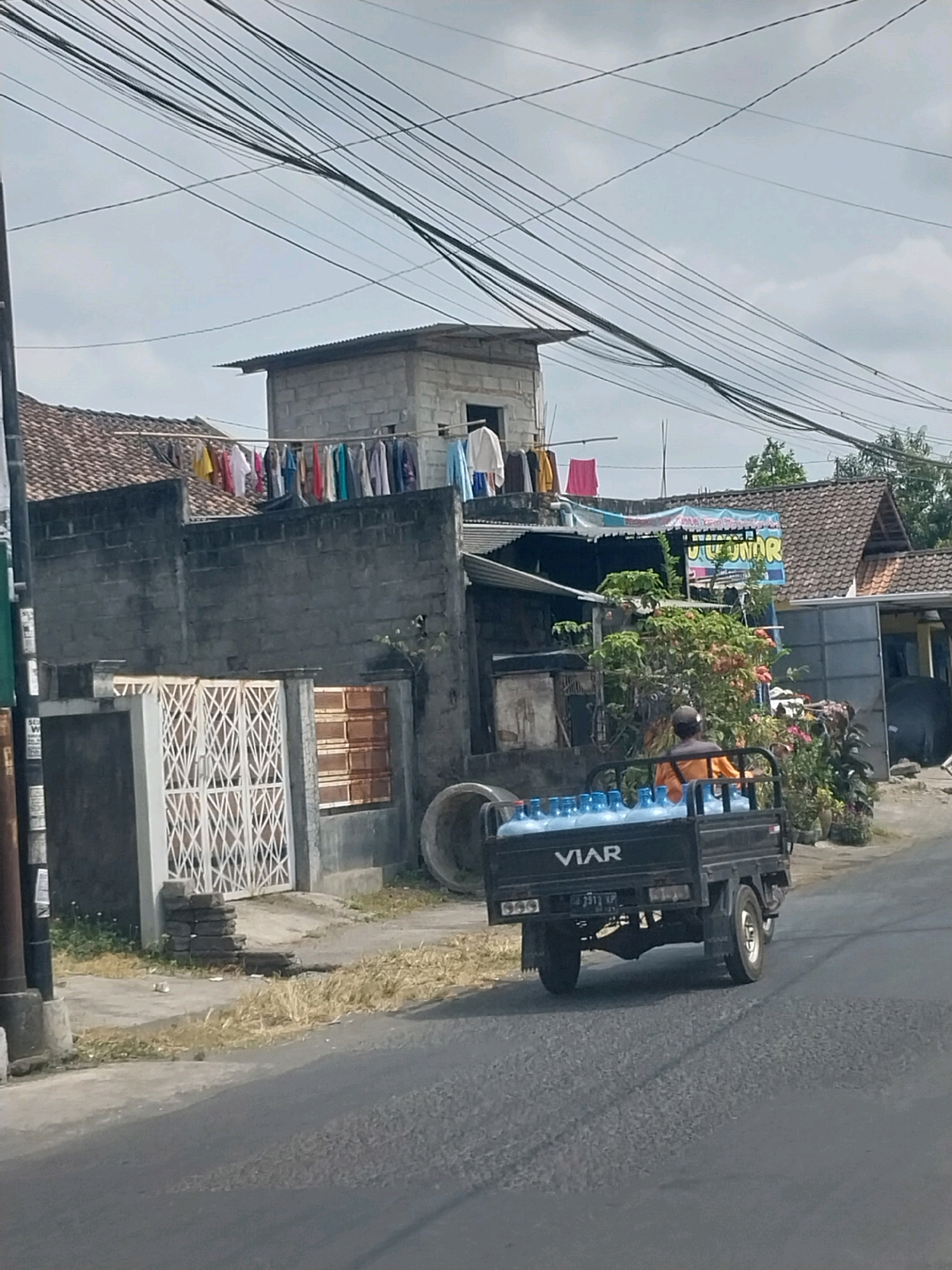 In the city of Purwomartani, Indonesia, a black VIAR pickup truck is seen driving on a paved road. The truck bed is loaded with multiple large blue water bottles. A person wearing an orange shirt and a dark cap is seated in the driver's seat. Behind the truck, a house with a tiled roof is visible. Laundry is hanging on a clothesline strung across the roof of the house. Further behind the house, more buildings and trees can be seen under a cloudy sky. The foreground shows a cracked asphalt road with dry grass and weeds growing along the side. A utility pole with many wires is also present on the left side of the image.