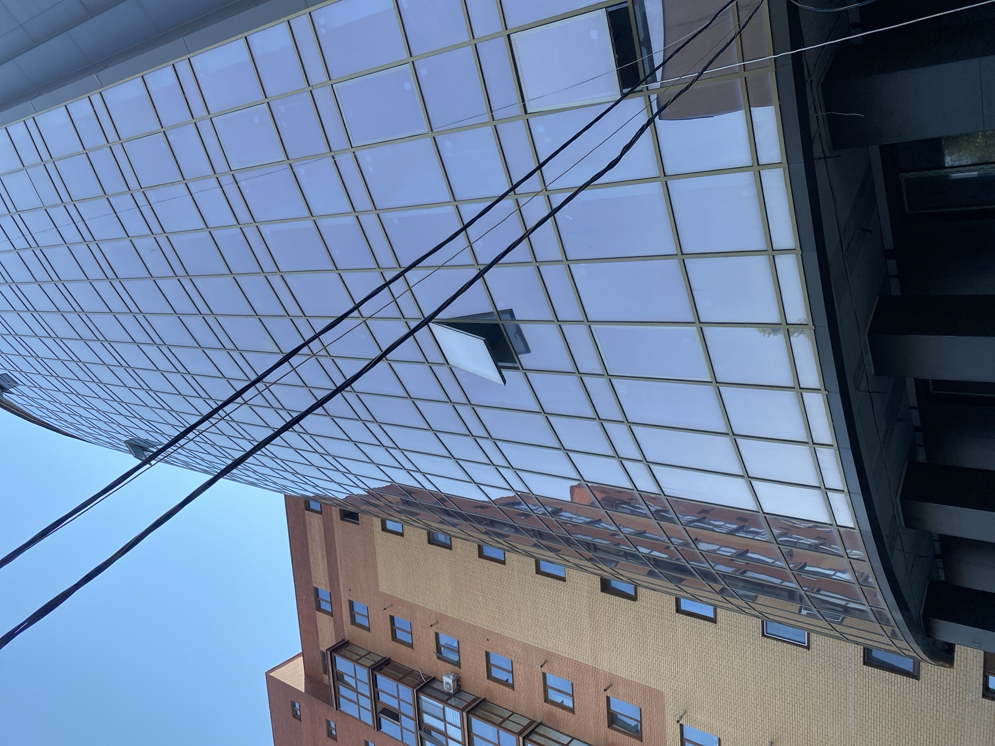 The image shows a low-angle view of two buildings against a clear blue sky. To the left is a brick building with a lighter beige section and red-brick sections, featuring multiple windows and balconies. To the right is a modern glass-facade building with a curved shape, composed of numerous square glass panels. Several dark power lines are strung across the foreground, obscuring parts of both buildings and the sky. One window on the glass building is open. The image appears to be taken in Vinnytsia, Ukraine, as indicated by the metadata.