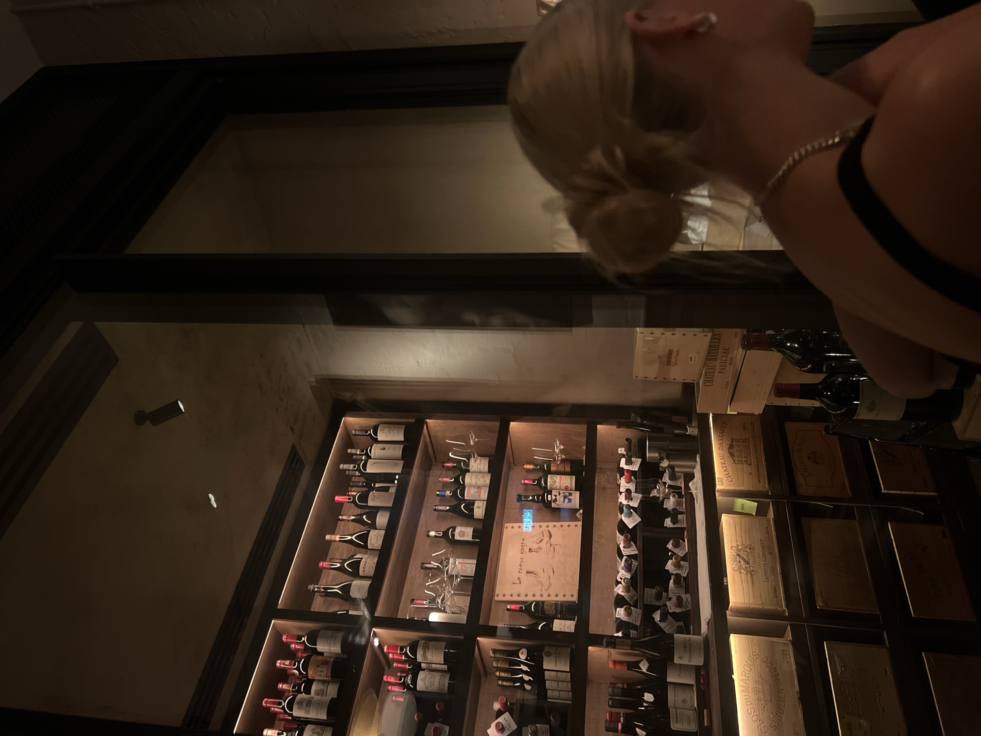 The image captures a low-lit, likely indoor scene focused on a wine display, partially obscured by a person.
A woman with blonde hair in a bun is partially visible in the upper right corner of the frame. The woman has a piece of jewelry on her neck and an earring on her right ear.
The primary subject is a tall, built-in wine rack, displaying numerous bottles of wine stacked in a grid-like pattern, including "CLOS DUMARQUIS," "CHATEAD," "LAFFITTE CARCASSET," "CHATEAU DASSAULT," and "PAUILLAC." There are also wooden crates.