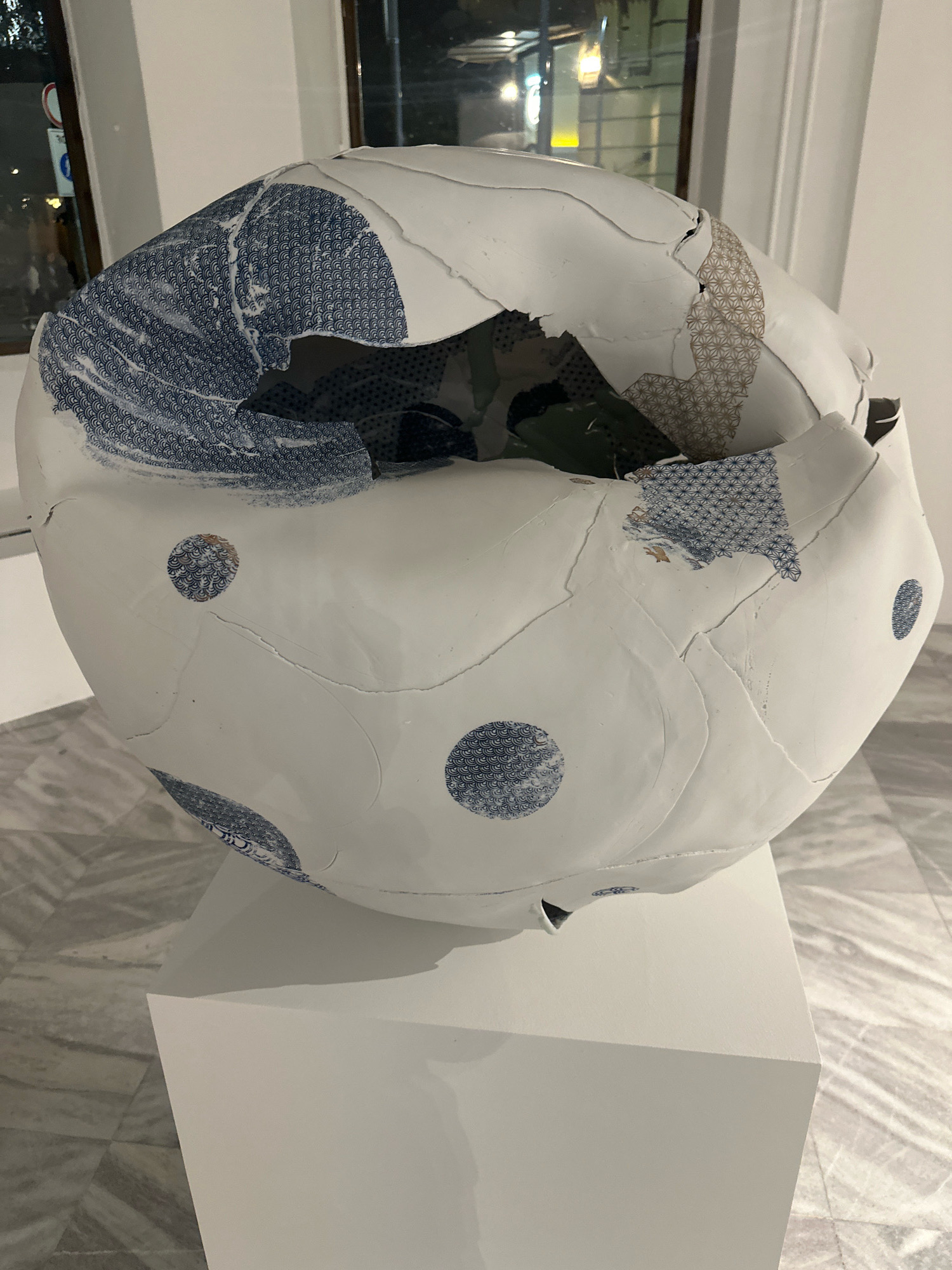 A large, broken, spherical ceramic art piece with white and blue patterned fragments sits on a white pedestal. The art piece has visible cracks and tears, revealing a dark interior. The exterior is decorated with blue circular patterns and a blue wave-like pattern. The background shows a window with reflections of a city at night and a marble floor. The art piece is located in Belgrade, Serbia.