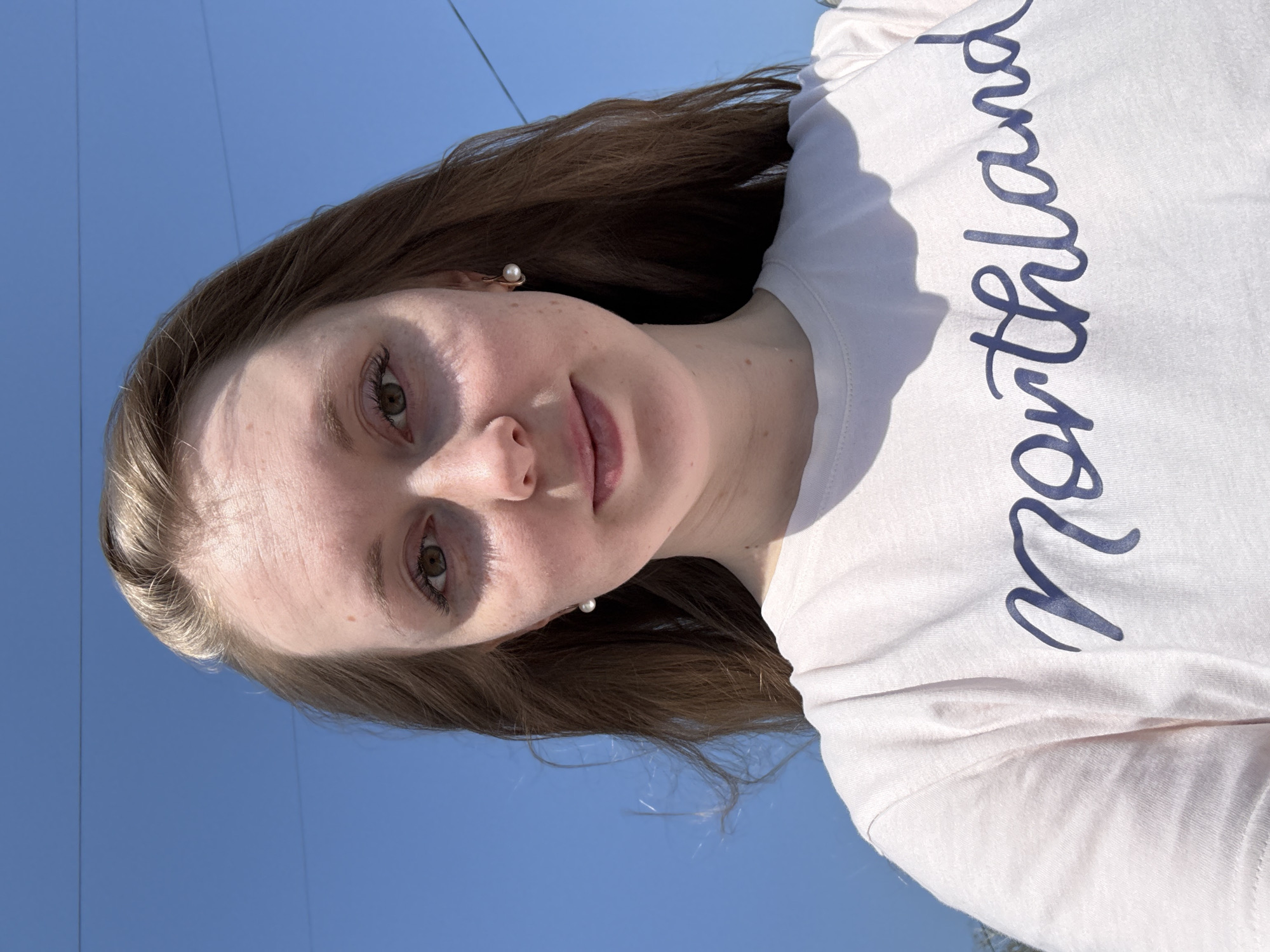 A woman is pictured in an outdoor selfie against a clear blue sky. She has fair skin, light brown hair, and is wearing pearl earrings. She is wearing a light-colored T-shirt with the word "Northland" printed on it in a dark cursive font. Fine lines from overhead wires can be seen in the top left corner of the frame. The location is Unknown, Unknown.