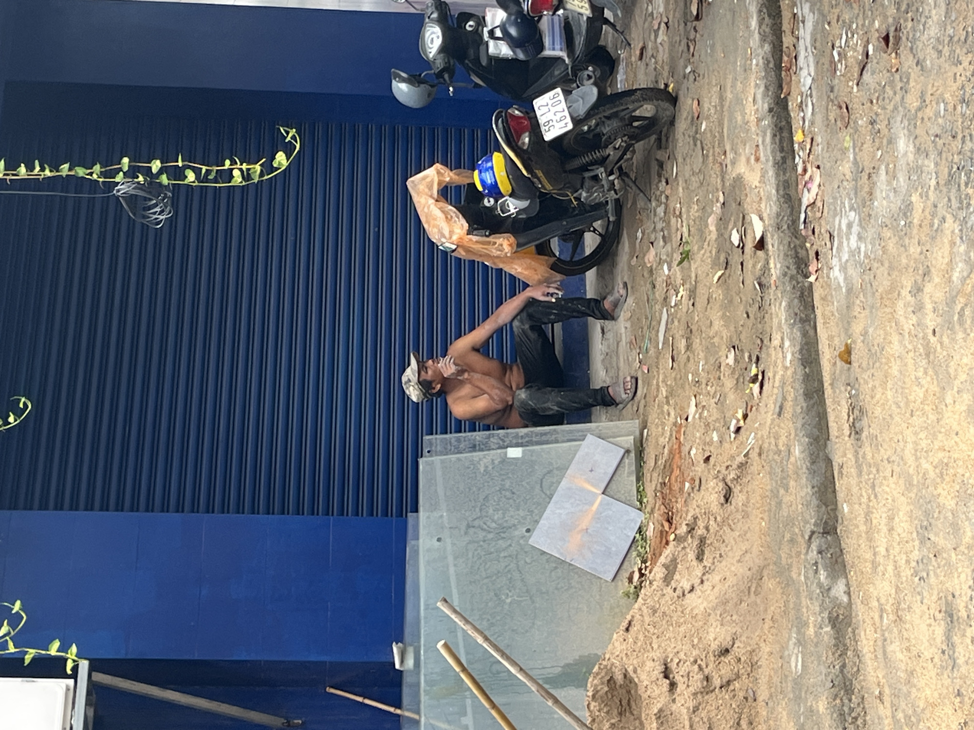 The image depicts a street scene in Nha Trang Ward, Vietnam. A bare-chested man in dark pants and a cap sits on the ground near a storefront with blue roll-up doors. A motorcycle is parked nearby, with a blue and yellow helmet hanging from the handlebars. Two square tiles lay on a glass sheet and some bamboo sticks are on the ground next to them. The sidewalk is concrete and littered with debris. Vines grow on the wall of the building.