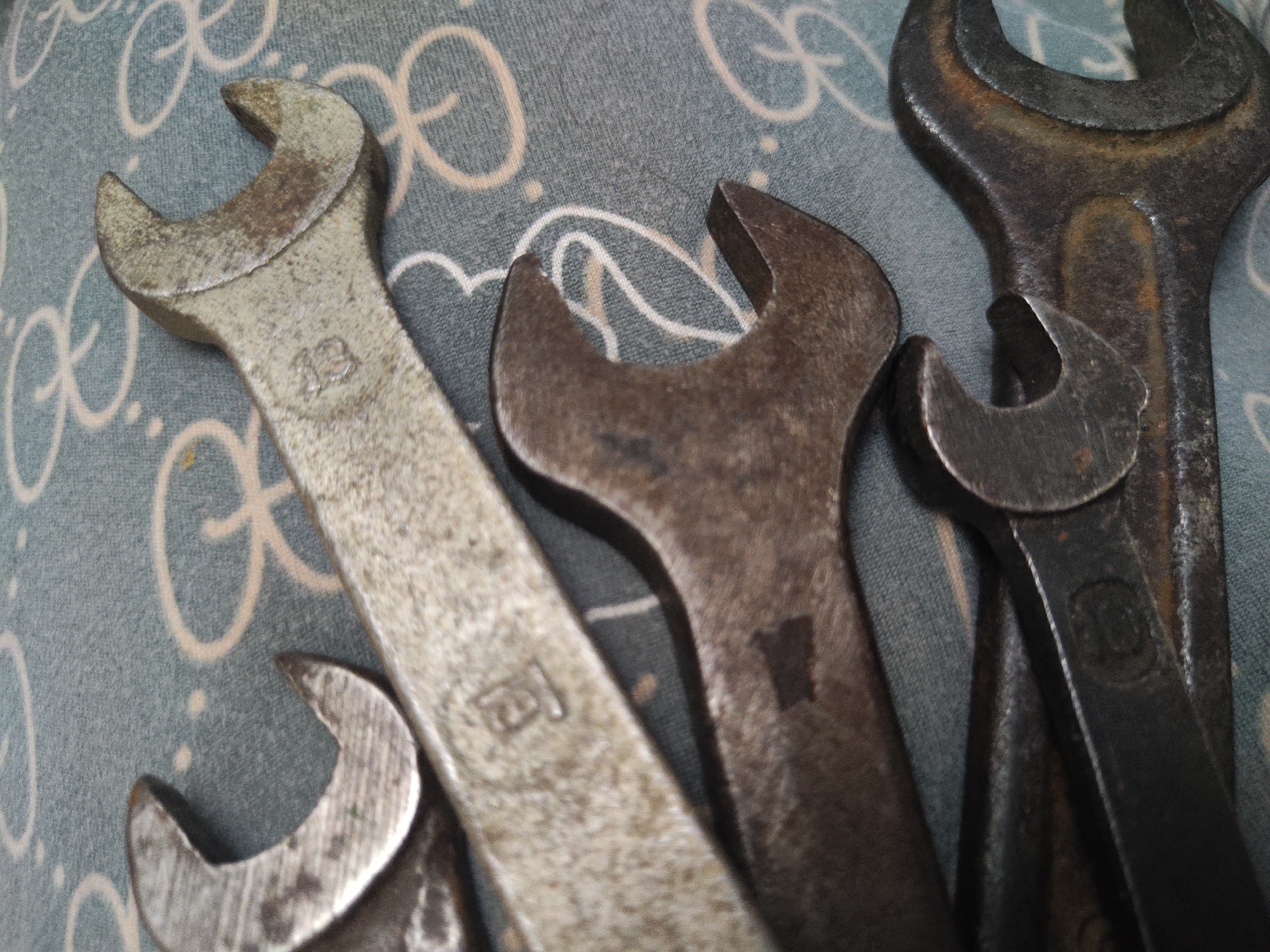 The image shows four open-end wrenches of various sizes lying on a surface with a patterned design. The wrenches are metallic, and show signs of wear and rust. The patterned surface appears to be a fabric, with a recurring circular or loop design. The tools and background are in focus, suggesting a close-up shot.
The location in the city of Pushkin, Russia cannot be determined from the image.