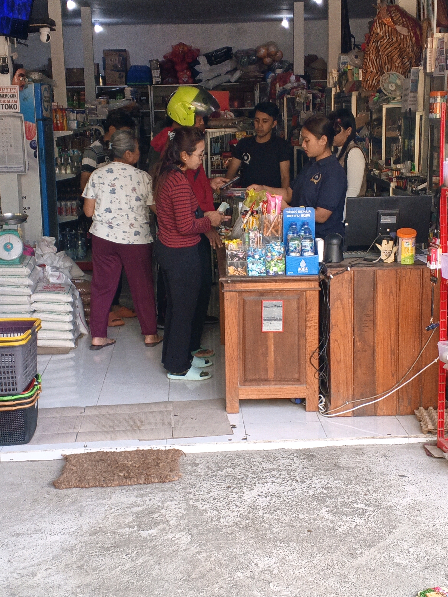 The image depicts the interior and entrance of a small retail store, likely a convenience store or warung, located in Penebel, Indonesia. The scene is bustling with activity. In the foreground, a brown coir doormat lies on a concrete walkway leading into the store.
Inside the store, several individuals are visible. To the left, a woman wearing a white floral shirt and maroon pants stands with her back to the camera, facing towards the interior. Next to her, a woman in a red and black striped shirt and black pants is engaged in a transaction, holding money in her hand. Behind her, a person wearing a yellow helmet and a red shirt is partially visible.
In the center of the store, a wooden counter with a glass display case filled with snacks and drinks is prominent. Two cashiers, a man in a black t-shirt and a woman in a navy blue uniform, are behind the counter attending to customers or stocking shelves. A computer monitor and various items are on the counter.
To the left of the counter, stacks of white sacks, likely containing rice or similar goods, are piled high, along with a green scale. A blue refrigerator is also visible. The shelves in the background are stocked with a variety of products, including food items, beverages, and household goods. The store appears to be well-organized and has a diverse inventory. The lighting suggests it is daytime, with overhead lights contributing to the illumination. The overall impression is of a busy local shop serving its community.