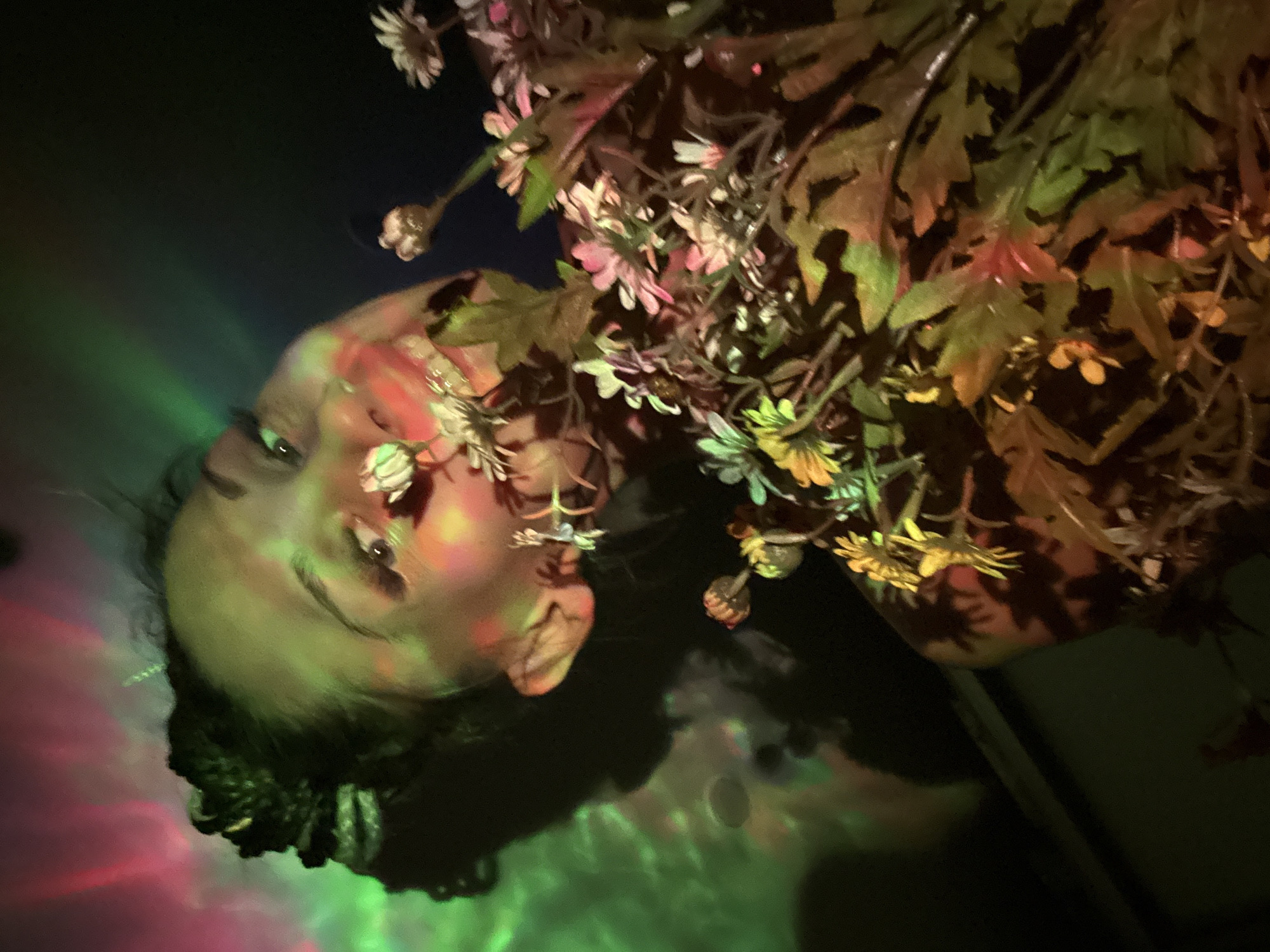 A person's face is visible, surrounded by various flowers and greenery. The lighting is peculiar, with green and pink hues casting shadows on the face and background. The subject's gaze is directed upwards, creating an ethereal effect. The presence of colorful lighting patterns suggests an indoor setting with artificial illumination. Location information is not present in the image.