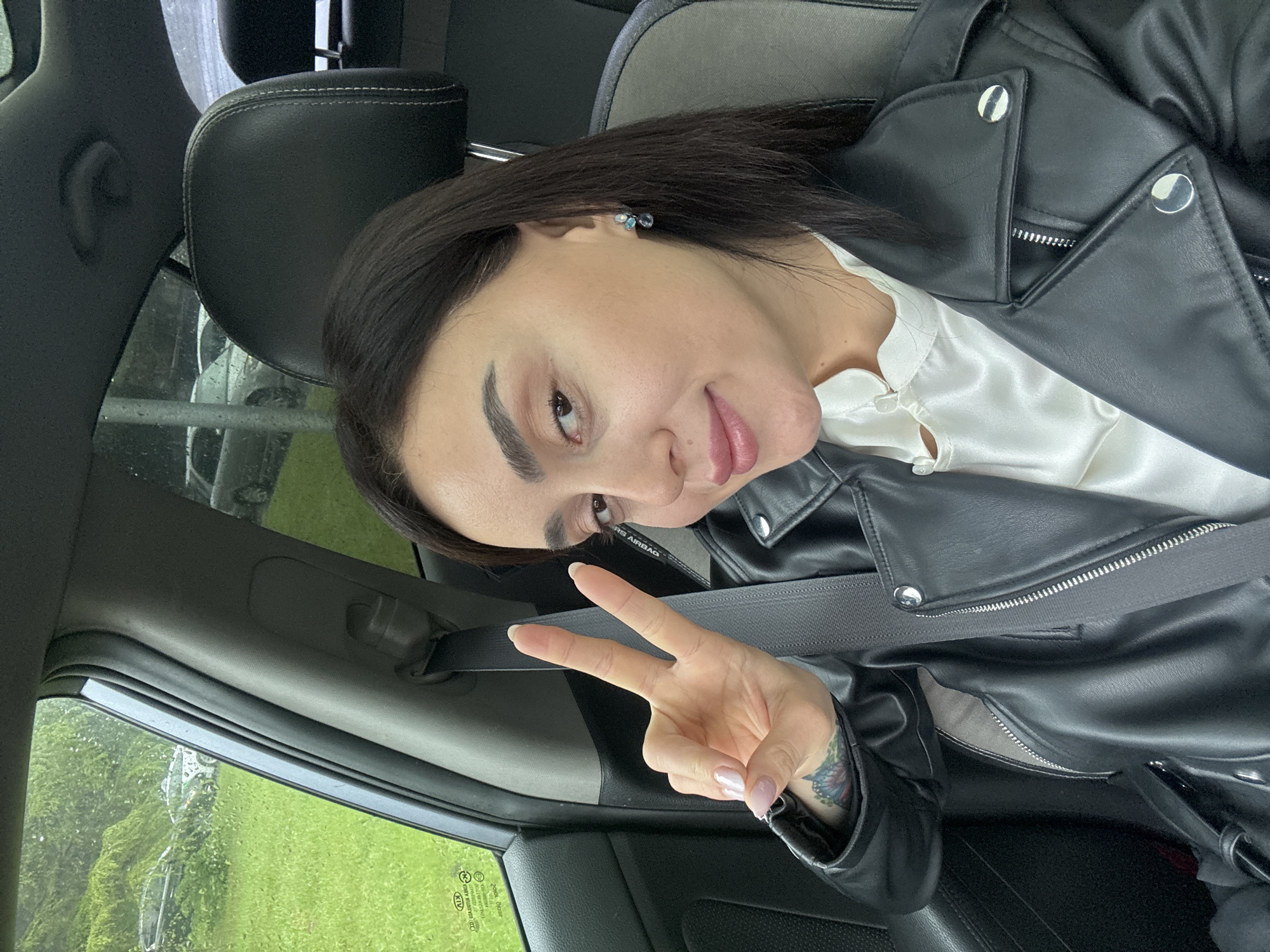 A woman is in a car, wearing a black leather jacket over a white top and a seatbelt. She is giving the peace sign with her right hand, which has a tattoo on the wrist. The car window shows a blurry green landscape, possibly indicating a rural or suburban area. The photo was taken during the daytime, with natural light illuminating the interior of the car. Without additional information, pinpointing the exact location in Tbilisi, Georgia, is not possible, as the surroundings are not specific to a particular urban area.