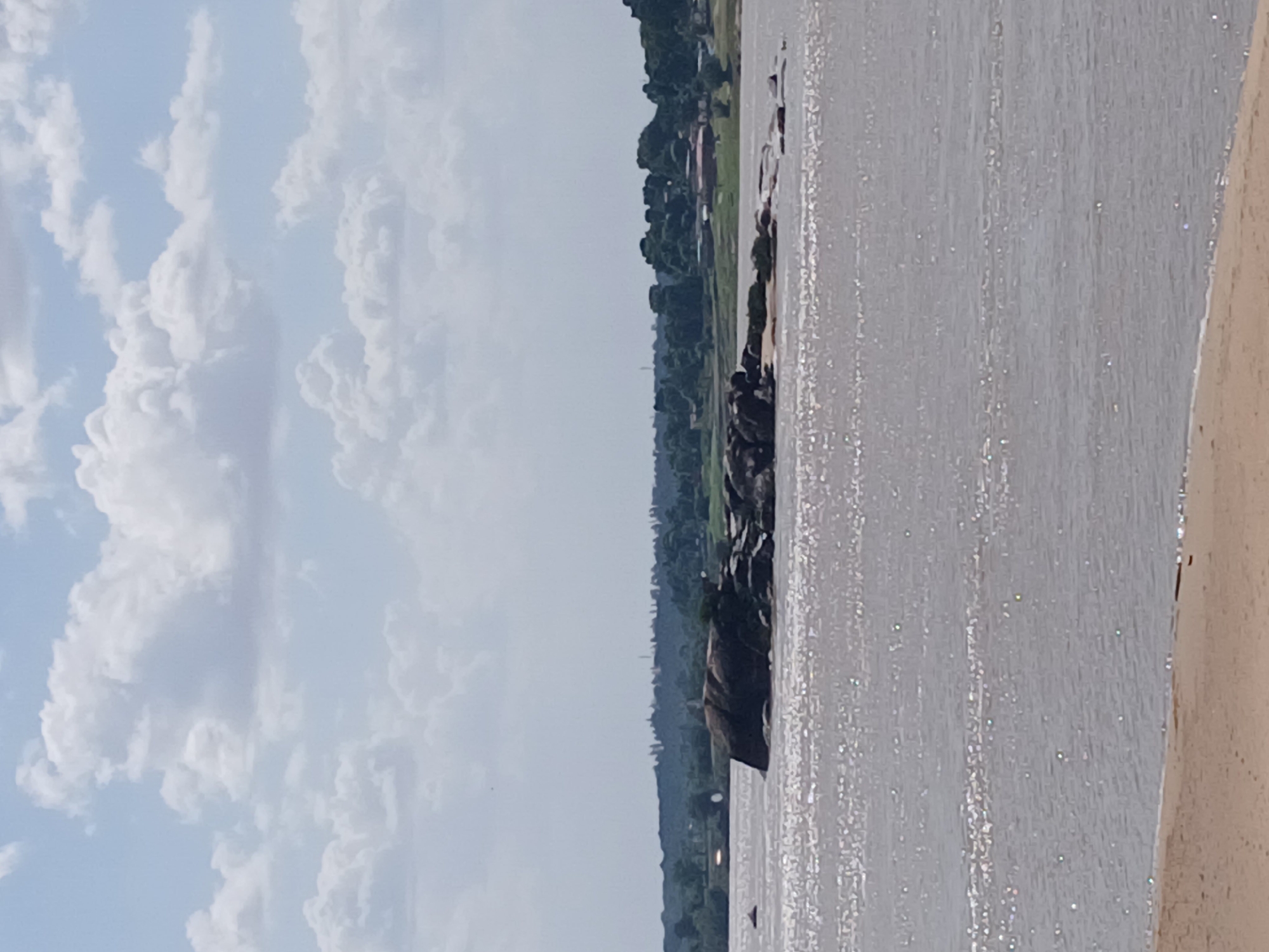 Here is a detailed description of the image:
The image depicts a body of water with visible reflections on the surface, likely a river or lake. Along the far bank, a dense line of trees and vegetation is visible, suggesting a natural or minimally developed area. Beyond the trees, hints of structures and settlements can be discerned. The sky is filled with scattered clouds on a mostly blue background. To the right of the water, there's an edge, potentially part of a structure like a wall or embankment. The overall scene appears to be an outdoor environment, possibly a waterfront area. The city and location is unknown.