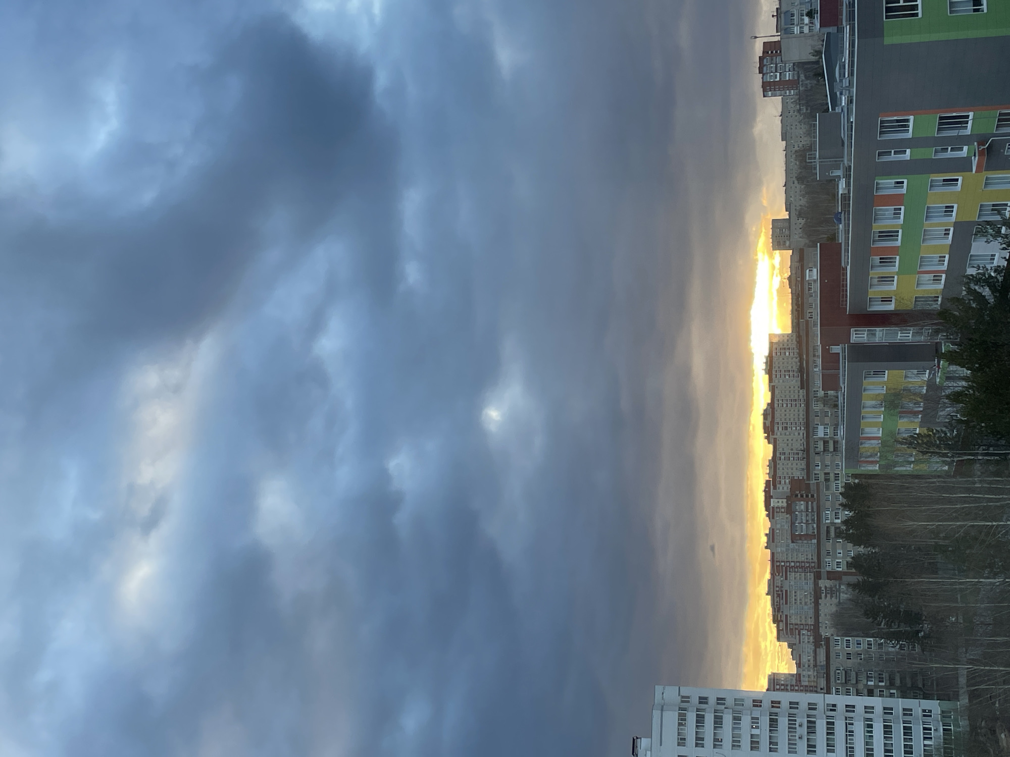 This panoramic image captures an urban landscape at either sunrise or sunset, characterized by a dramatic sky and dense residential architecture. The horizon is bisected by an intense, brilliant band of golden and yellow light, indicating the low position of the sun. Above this luminous strip, the sky is dominated by expansive, heavy, dark grey clouds, creating a striking contrast between the bright horizon and the somber, overcast atmosphere. Patches of lighter blue and white are visible within the cloud cover, particularly towards the upper left.
The scene is filled with numerous multi-story apartment buildings, typical of cities like Izhevsk, Russia. These structures vary in design, with some appearing older and more uniform, while others showcase modern, colorful facades featuring panels in vibrant greens, yellows, oranges, and various shades of grey and white. Many windows are visible across the building exteriors.
In the mid-ground, a line of bare trees, likely deciduous, stands between the buildings, suggesting a late autumn or winter setting. The perspective is an elevated one, possibly taken from a high floor of another building, offering a wide view over the sprawling city. There is no discernible human activity or visible text within the frame. The image emphasizes the interplay of natural light and urban structures under a dramatic sky.
