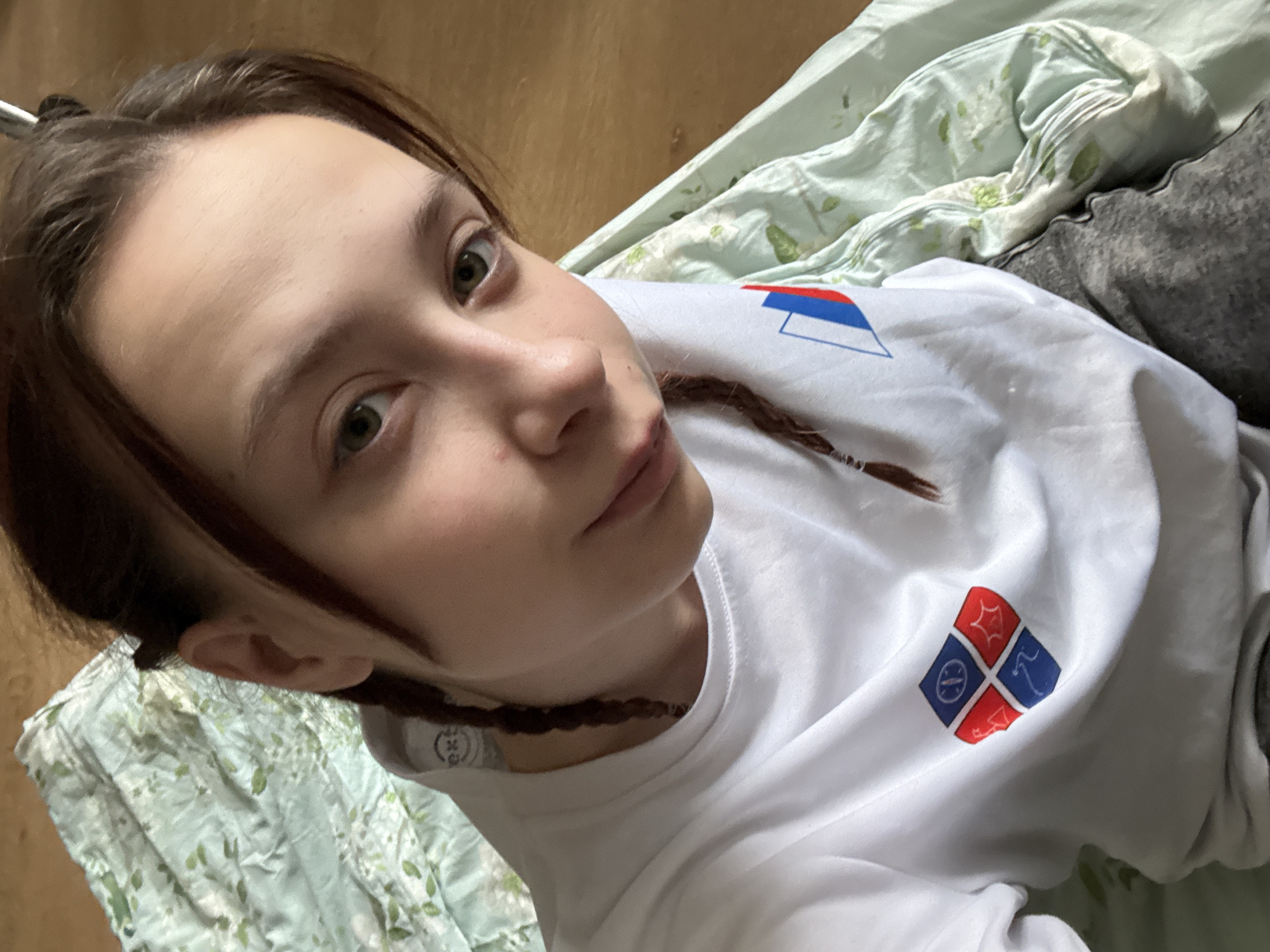 A young woman lies on what appears to be a bed. She wears a white long-sleeved shirt with two insignia near her shoulders: one consisting of blue, white, and red horizontal stripes, and another a diamond shape divided into quadrants containing various symbols. Her brown hair is braided, and she looks directly at the camera. She is lying on a floral-patterned bedspread with a gray garment on top. The background includes a wooden floor. This image was taken in Berezniki, Russia.