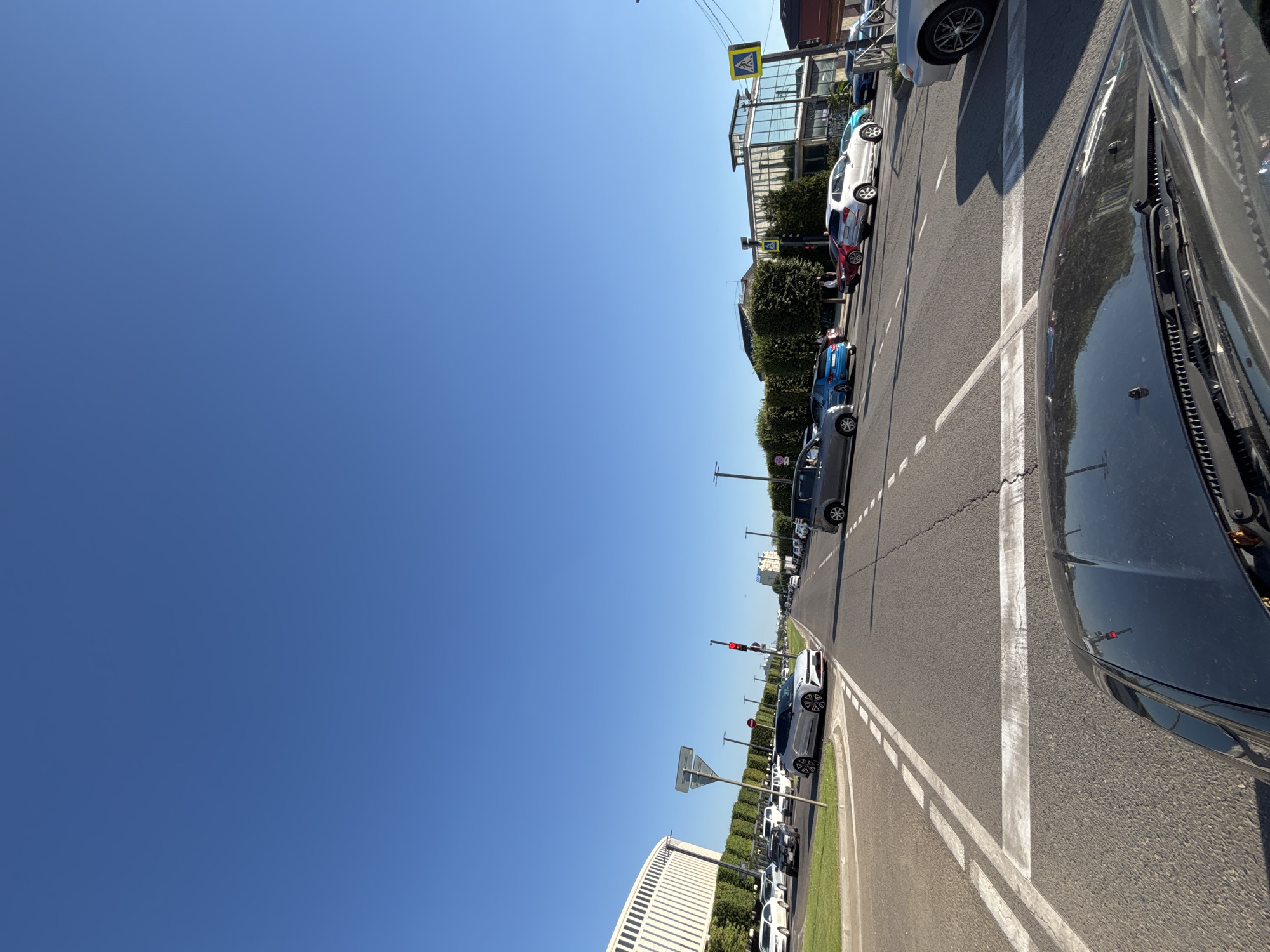 This is a media file showing a street scene in Krasnodar, Russia. The image is taken from inside a vehicle, looking out at a street with multiple lanes of traffic. Several cars are parked along the side of the road, and others are driving in the same direction. The street is lined with trees and buildings. The sky is clear and blue, suggesting a sunny day. There are pedestrian crossings visible, indicated by white lines on the road and signage. Traffic lights are also present, with one showing a red light. The overall scene depicts a typical urban environment during daytime.