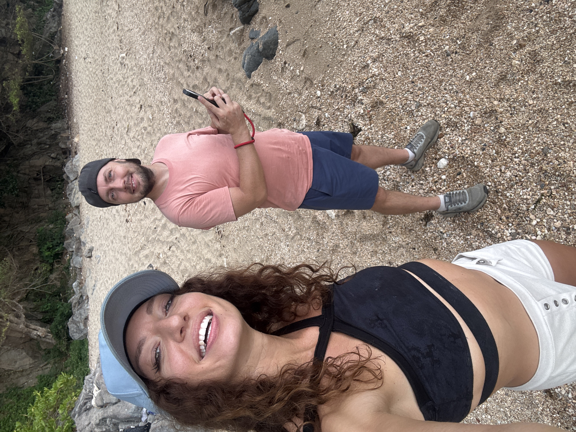 Two fair-skinned individuals pose for a selfie at a beach, potentially in Đà Nẵng, Vietnam. The woman, positioned closer to the camera, wears a light blue baseball cap, a black multi-strap top, and white shorts. She has curly brown hair and is smiling broadly. Behind her, a man stands in a light pink t-shirt, blue shorts, grey sneakers, and a dark grey baseball cap. He is holding a smartphone in his hands. The ground is composed of sand and small pebbles, with some rocky terrain visible near the edges of the frame. Vegetation is present on the left side of the image.